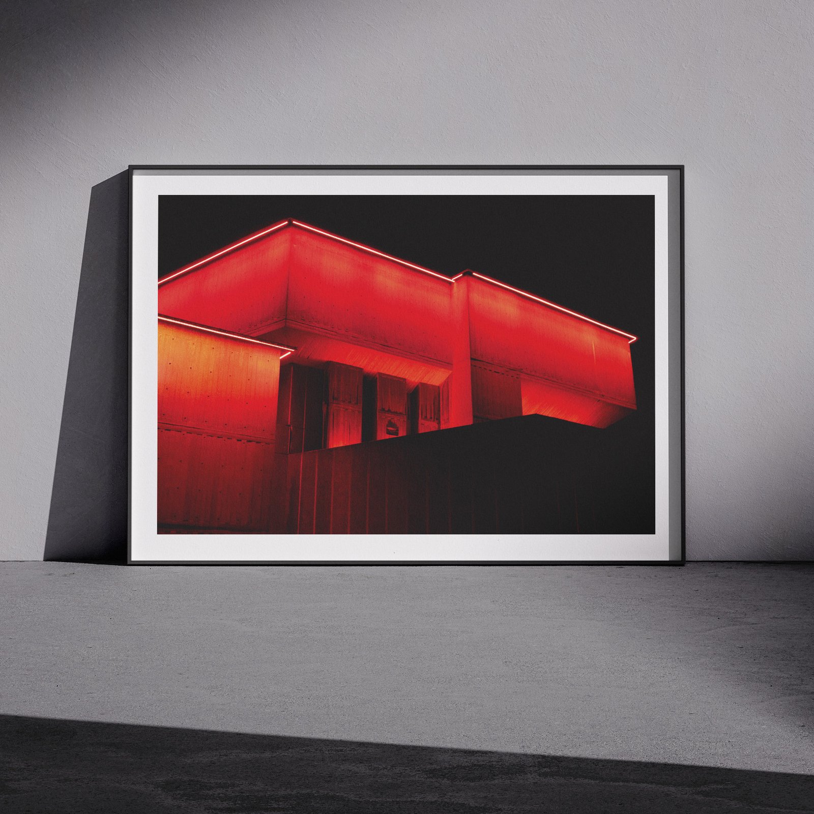 Horizontal architectural photograph of a neon-glowing red building structure in a white frame with black outer frame, leaning against a pale wall in a gallery-style floor presentation with soft studio lighting creating subtle shadows.