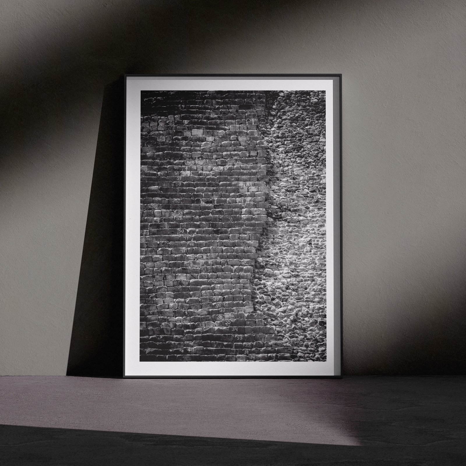 Vertical black and white architectural photograph of a brick or stone wall texture with dramatic directional light creating strong relief detail, displayed in a white frame with white mat, leaning against a dark charcoal wall in a gallery-style floor presentation with dramatic side lighting.