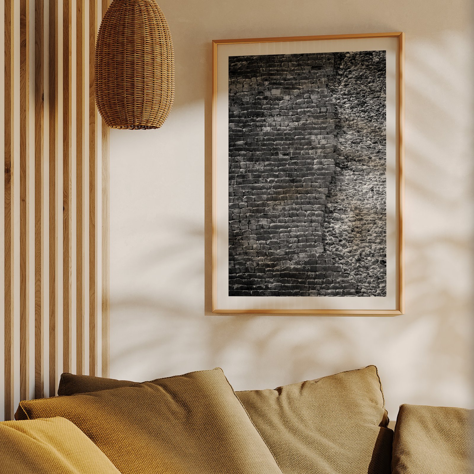 Vertical black and white textural photograph of layered brick or masonry surface in a natural wood frame with white mat, hung on a cream wall above a mustard-gold upholstered sofa, styled with wooden slat wall paneling and a woven rattan pendant light visible above.