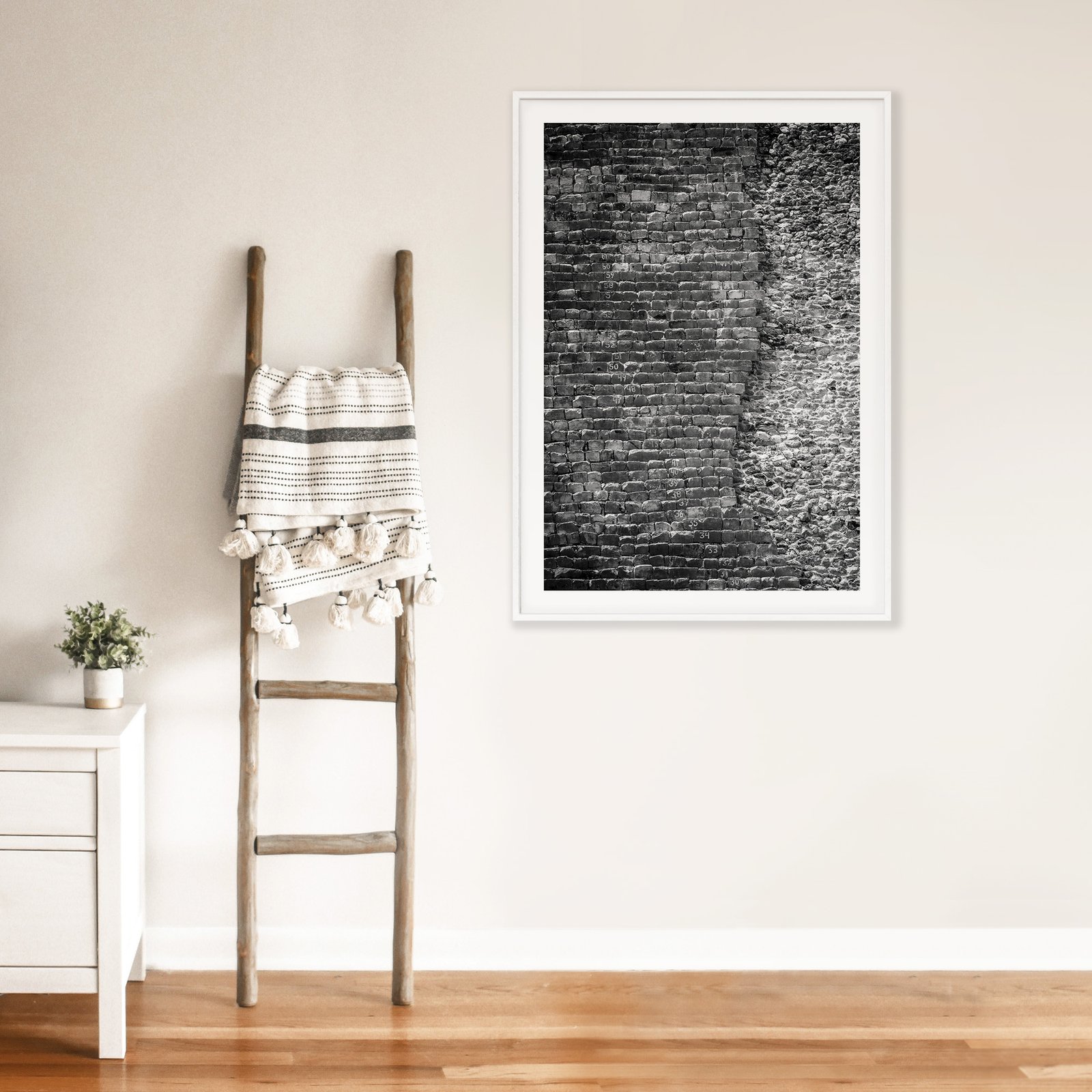 Vertical black and white architectural texture photograph of a brick wall with dimensional surface detail in a white frame with white mat, hung on a cream wall above a wooden ladder with rolled linens, styled with a small white side table displaying a potted plant on warm wood flooring.