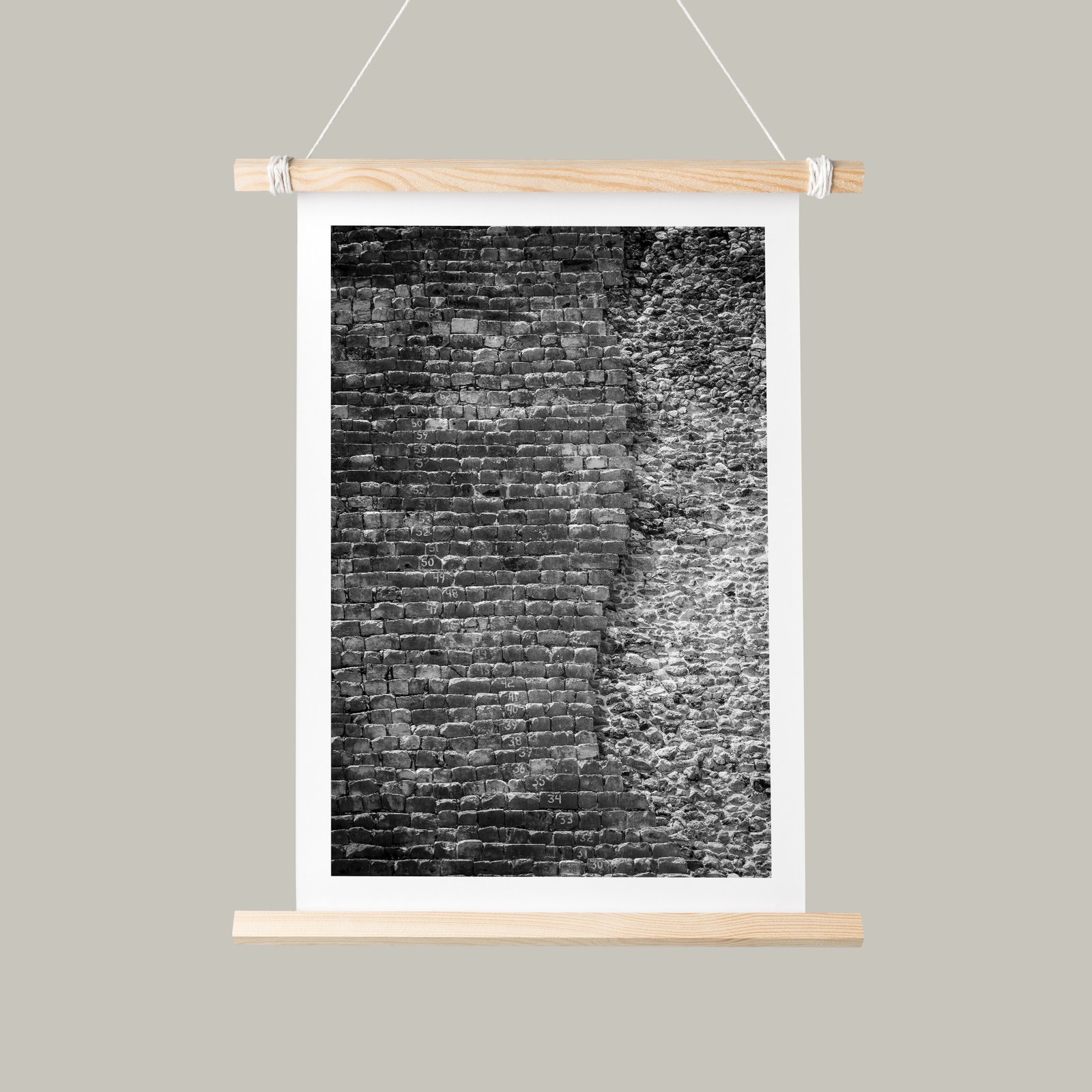 Vertical black and white textural brick wall photograph mounted as a hanging banner with white mat, natural wood dowel top and bottom frame with rope suspension, displayed against a soft sage-green background.