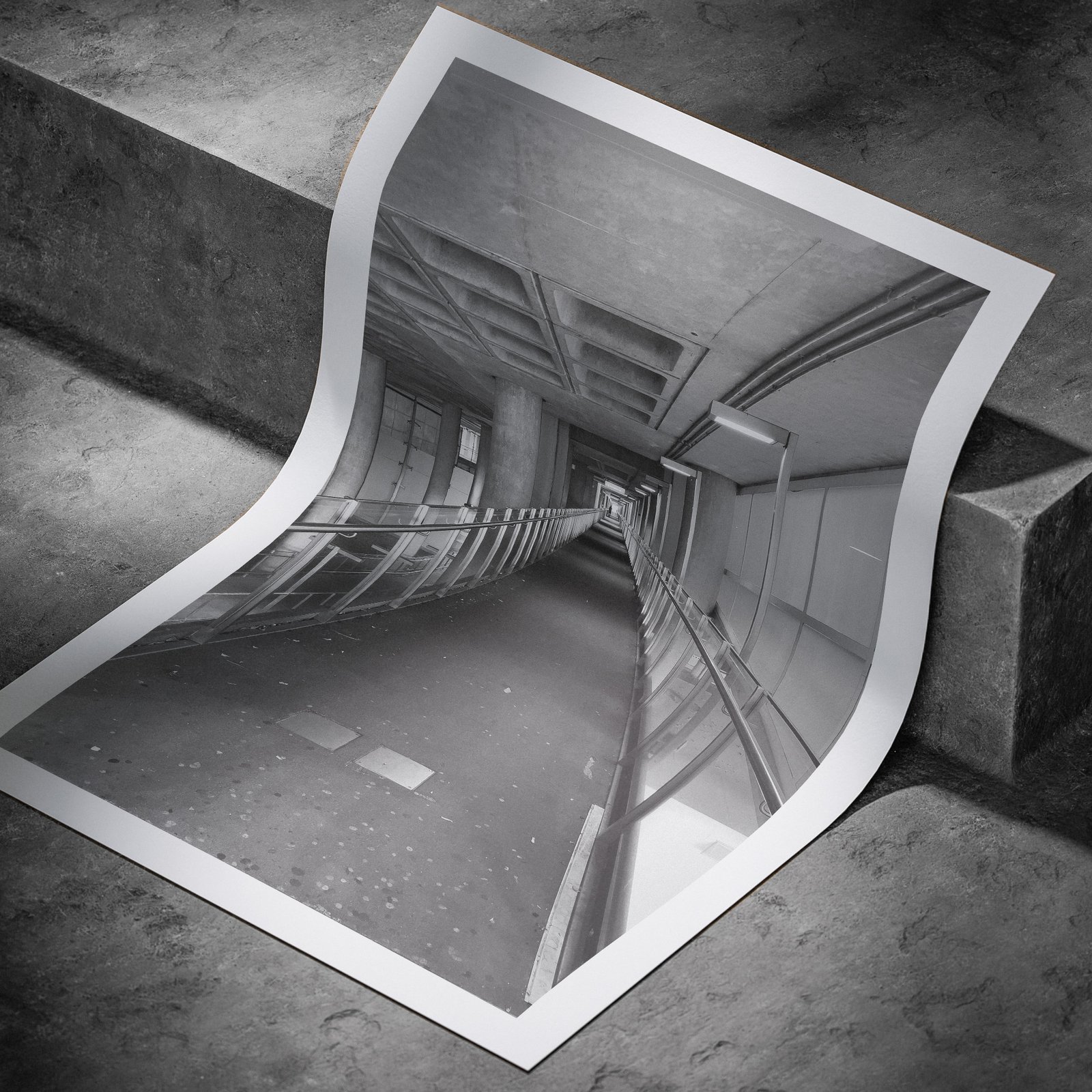 Vertical monochromatic architectural photograph of a corridor interior with perspective lines and railings printed on white paper, displayed in a curved, rolled perspective on a concrete surface with dramatic angled lighting highlighting the print edges and linear composition.