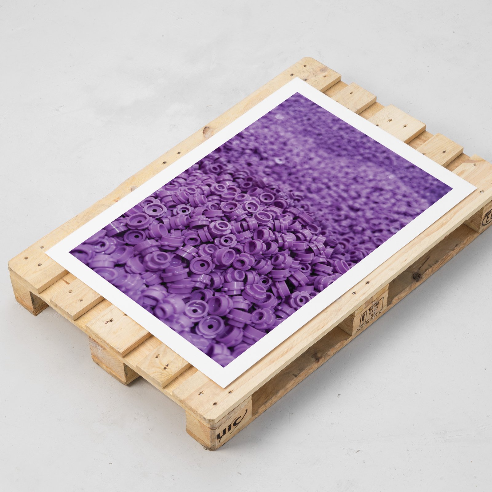 Vertical aerial photograph of a dense arrangement of purple LEGO bricks viewed from above, creating a textured pattern of studs and interlocking blocks, displayed on a natural wood stretcher-frame pallet with visible mounting hardware, shown at an isometric perspective angle on a light background, emphasizing the three-dimensional presentation.