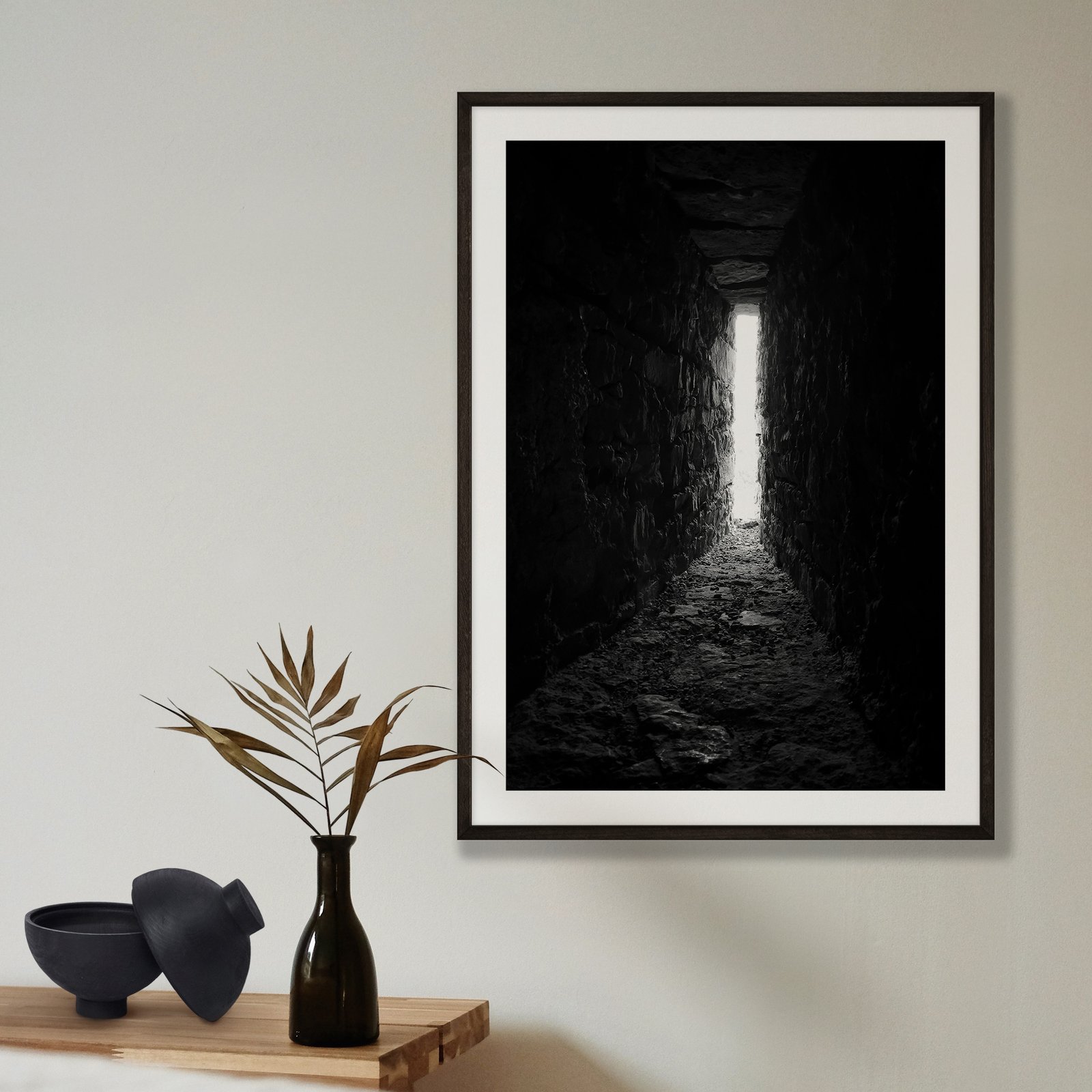 Vertical black and white architectural photograph of a dramatic cave or tunnel interior with dark rough stone walls and a bright opening creating a strong silhouette effect and focal point of light, displayed in a black frame with white mat on a pale beige wall, styled with a minimalist shelf arrangement below featuring a black vase with dried grasses and black ceramic bowl.