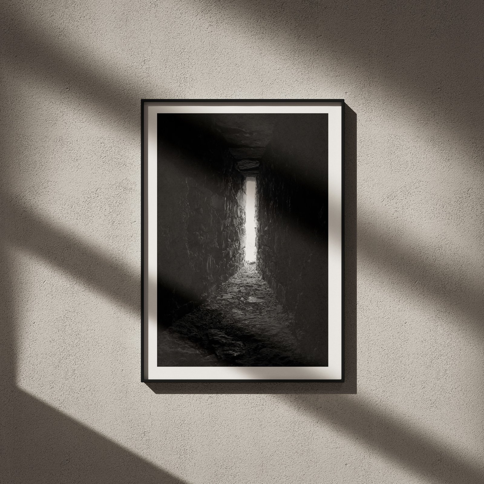 Vertical black and white cave or tunnel interior photograph with dramatic light entering from the opening in a black frame with white mat, hung on a neutral wall with strong directional window light creating dramatic geometric shadows across the entire composition.