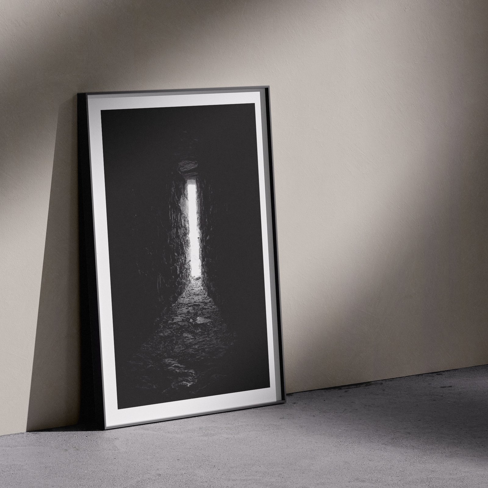 Vertical black and white architectural cave interior photograph showing luminous opening and textured stone walls in a white frame with white mat, leaning against a pale wall in a gallery-style floor presentation with soft studio lighting creating subtle shadows.