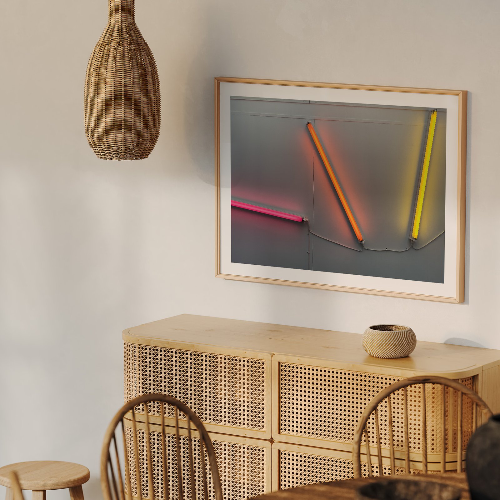 Horizontal neon art photograph featuring glowing pink, orange, and yellow tubes creating an abstract composition in a natural wood frame, hung on a cream wall above a natural wood dresser with rattan details, styled with a woven rattan pendant light above and wooden chair visible below.