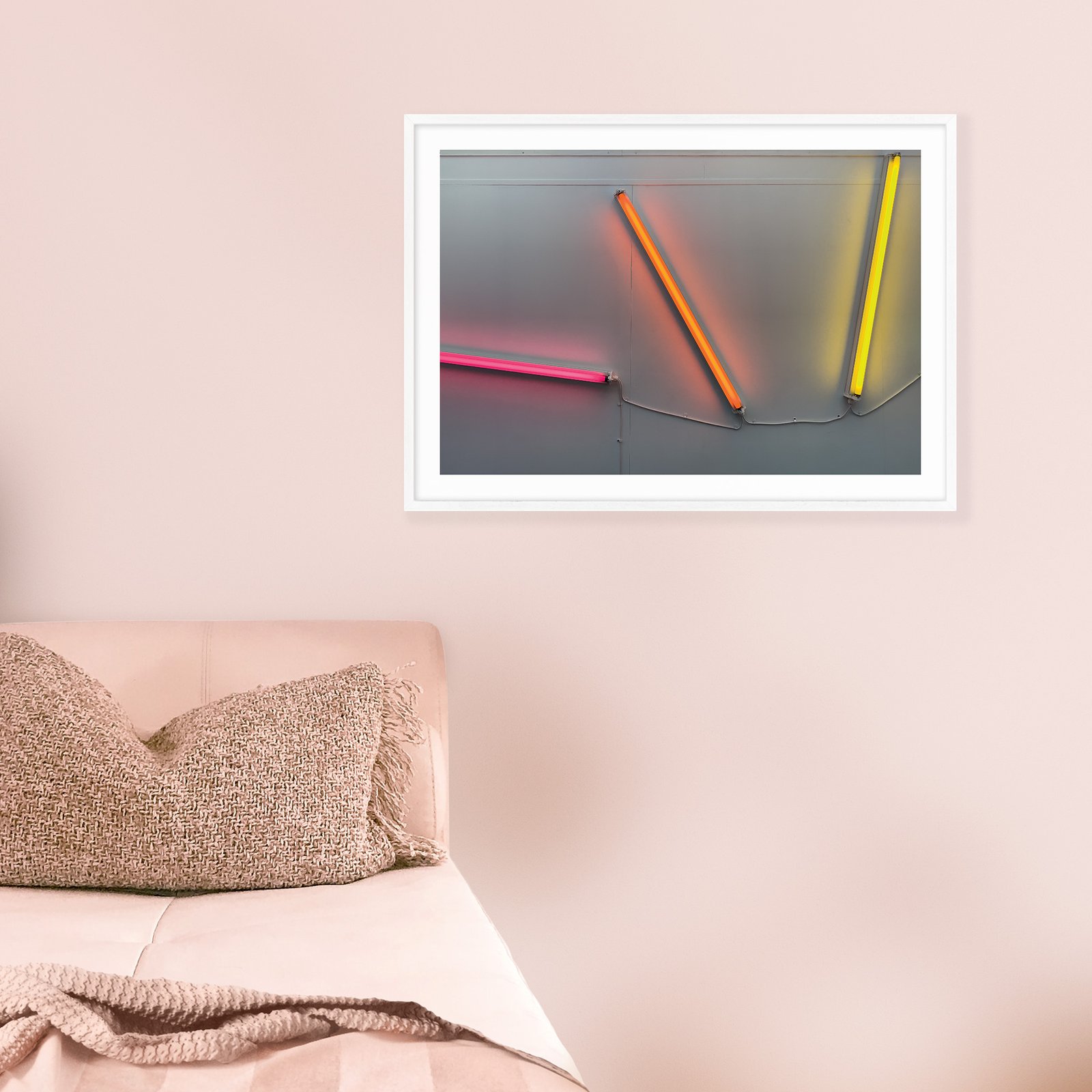 Horizontal contemporary neon light artwork with vibrant pink, orange, and yellow luminous tubes in a white frame with white mat, hung on a soft dusty pink wall above a bed with textured cushions and neutral bedding, creating a vibrant bedroom focal point.