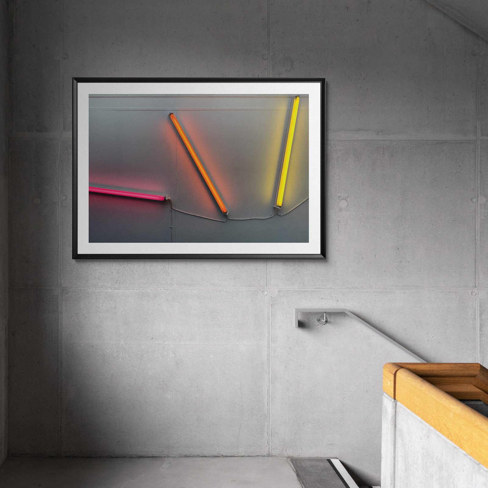 Horizontal neon art photograph showing glowing pink, orange, and yellow tube arrangement in a black frame with white mat, hung on a textured dark gray industrial wall with concrete finish and steel hardware visible nearby.
