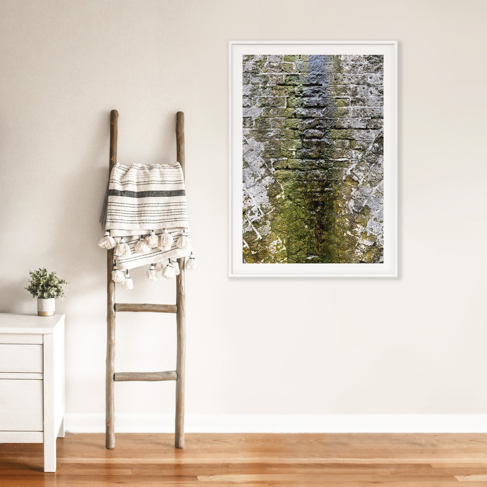 Vertical architectural close-up photograph of a weathered railway arch covered in green moss and lichen with gray stone texture, displayed in a white frame with white mat, hung on a cream wall above a wooden ladder with rolled linens, styled with a small white side table displaying a potted plant on warm wood flooring.