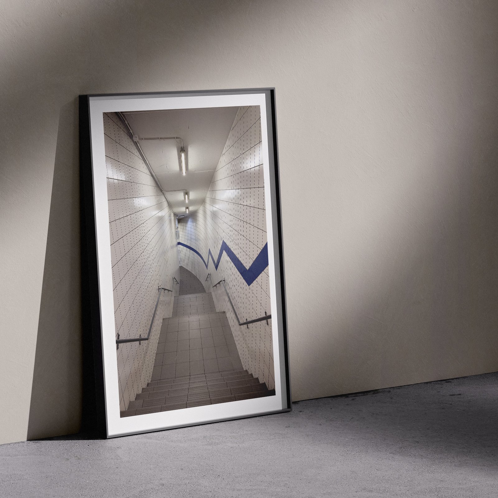 Vertical architectural photograph of a contemporary car park staircase with fluorescent lighting, white tile surfaces, and blue arrow markers in a white frame, leaning against a pale wall in a gallery-style floor presentation with soft studio lighting creating subtle shadows.