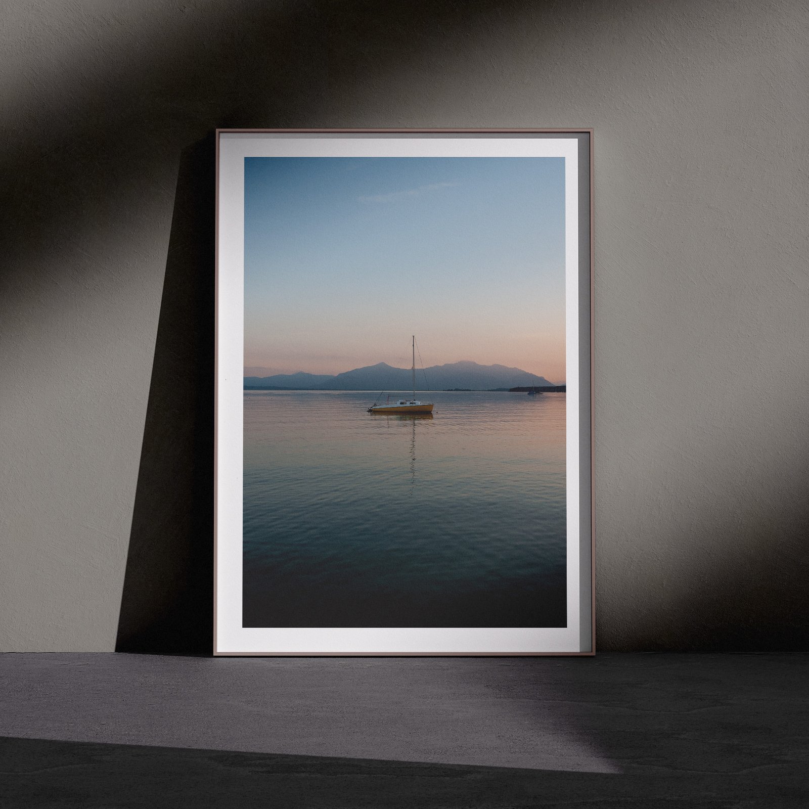 Vertical seascape photograph of a lone sailboat on still water with soft sky tones in a white frame, leaning against a dark charcoal wall in a gallery-style floor presentation with dramatic side lighting.