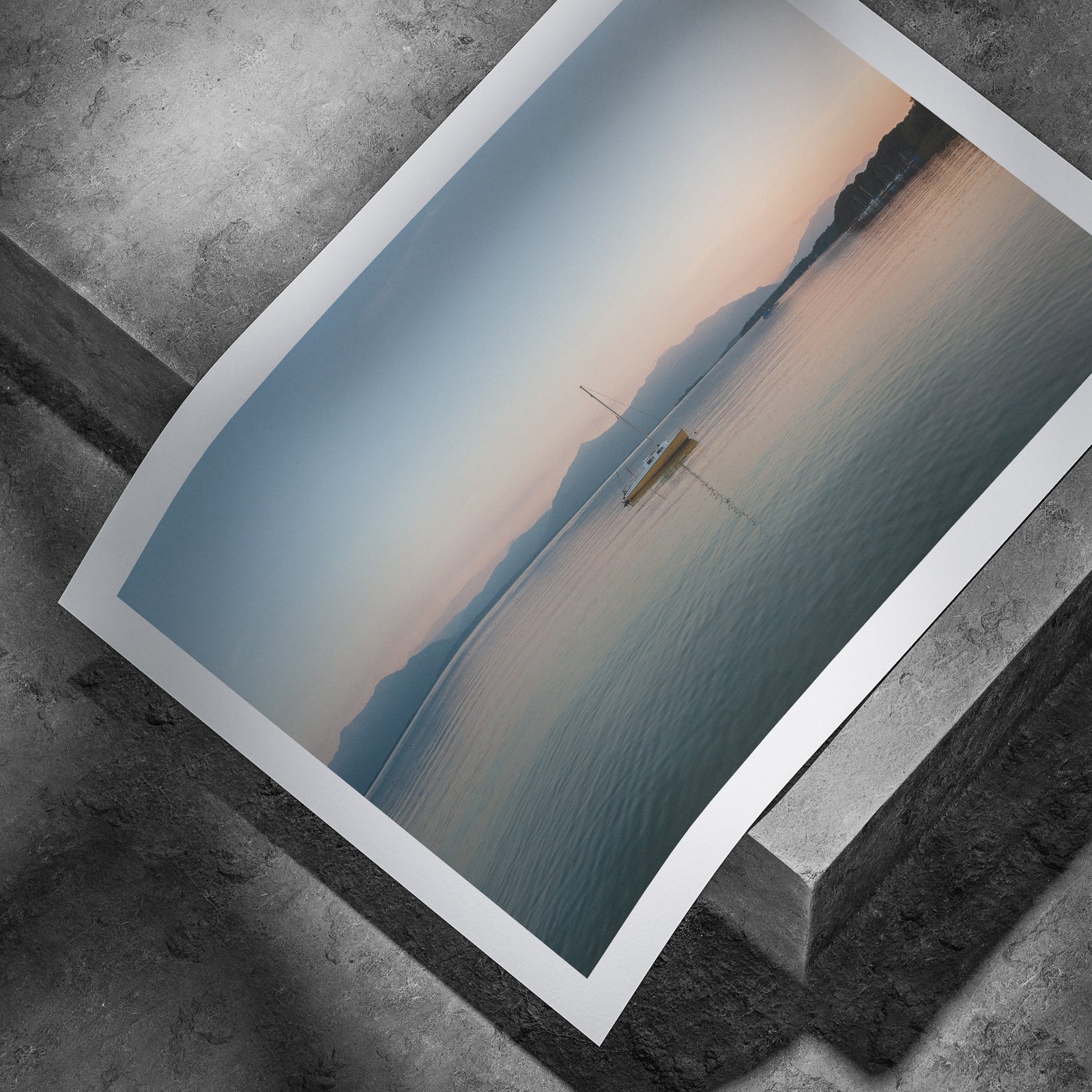 Horizontal seascape photograph of a peaceful sailboat scene in calm water printed on white paper, displayed in a curved, rolled perspective on a concrete surface with dramatic angled lighting highlighting the print edges.