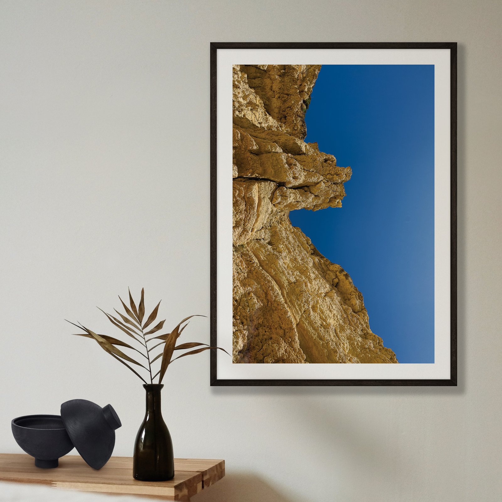 Vertical architectural photograph of a golden-tan rocky cliff formation with dramatic weathered texture against a clear bright blue sky, displayed in a black frame with white mat on a pale beige wall, styled with a minimalist shelf arrangement below featuring a black vase with dried grasses and black ceramic bowl.