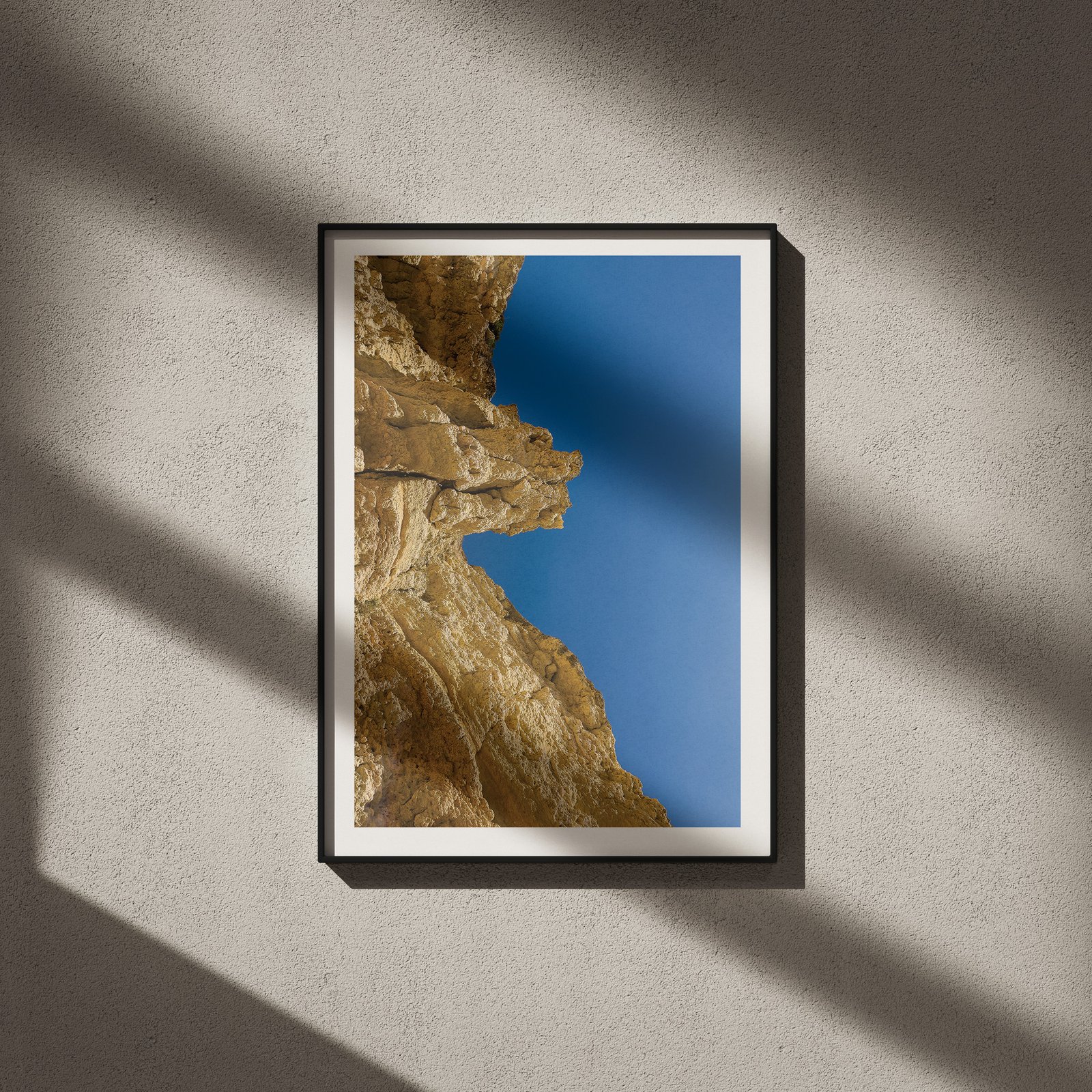 Vertical architectural photograph of a golden-tan rocky cliff formation with clear blue sky in a black frame with white mat, hung on a neutral wall with strong directional window light creating dramatic geometric shadows across the entire composition.