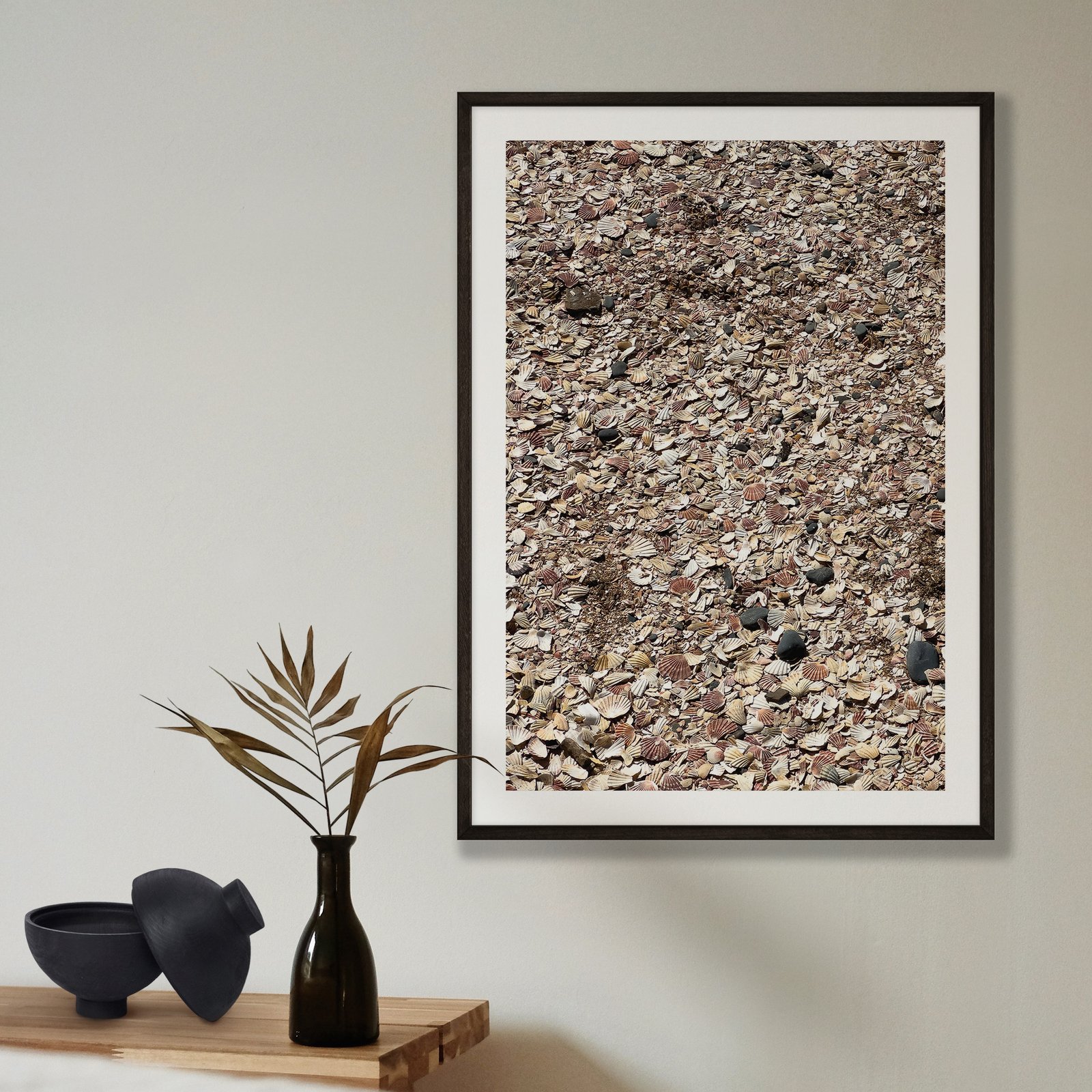 Vertical abstract photograph of natural gravel and stone texture with warm tan and gray tones in a black frame with white mat, hung on a pale beige wall, styled with a minimalist shelf arrangement below featuring a black vase with dried grasses and black ceramic bowl.