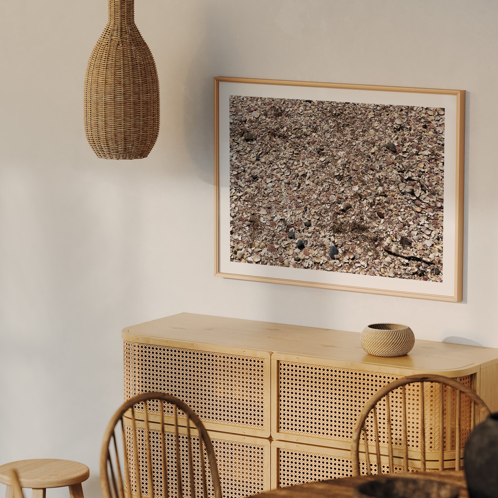 Vertical abstract textural photograph of natural stone and gravel in a natural wood frame with white mat, hung on a cream wall above a natural wood dresser with rattan details, styled with wooden chairs visible below.