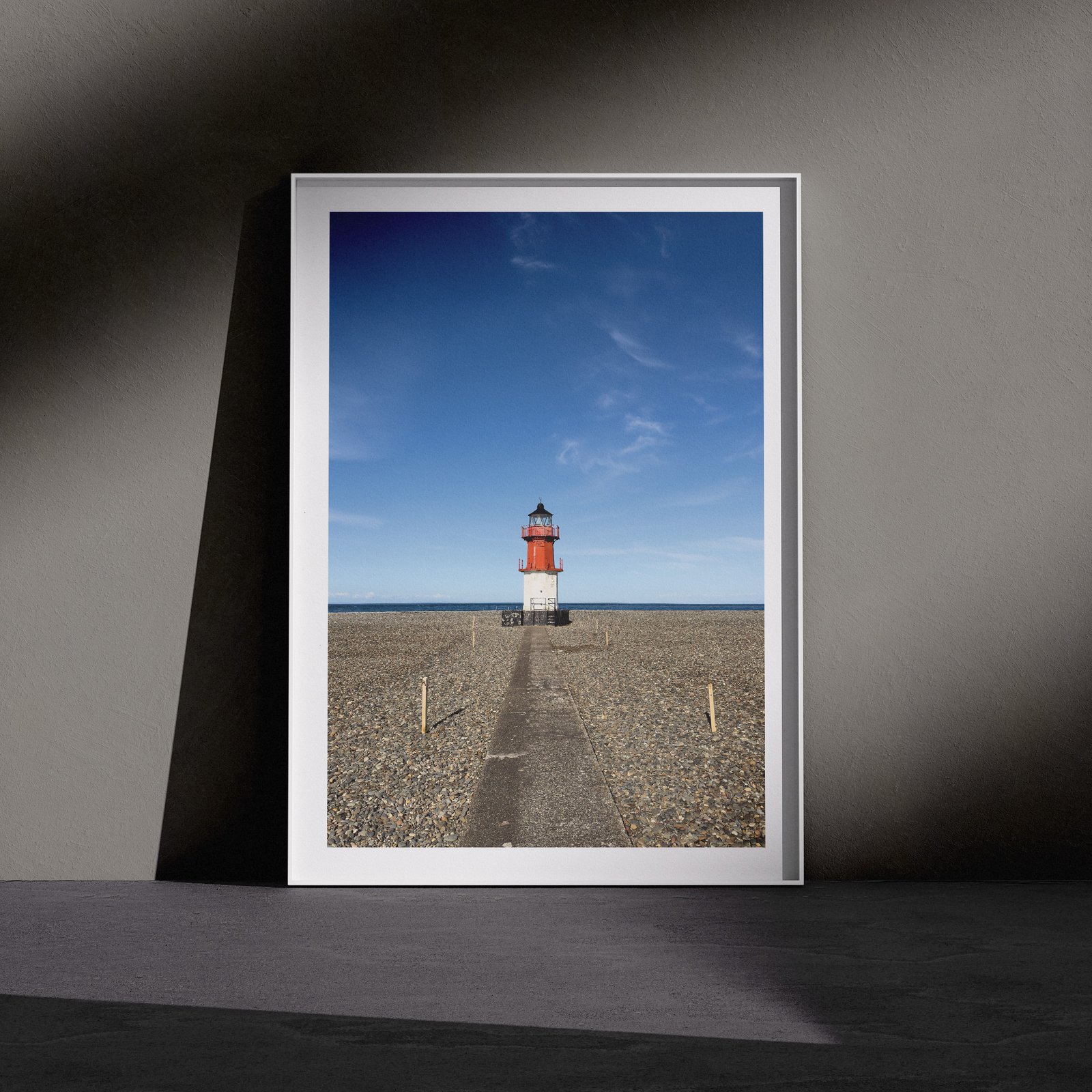 Vertical landscape photograph of a red and white striped lighthouse standing solitary on expansive tidal mudflats beneath clear blue sky, displayed in a white frame with white mat, leaning against a dark charcoal wall in a gallery-style floor presentation with dramatic side lighting.