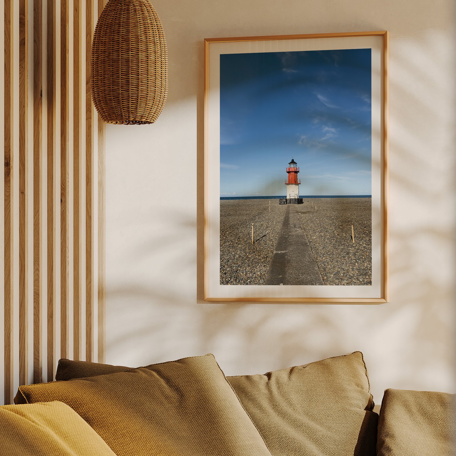 Vertical landscape photograph of a minimalist lighthouse on flat mudflats with clear sky in a natural wood frame with white mat, hung on a cream wall above a mustard-gold upholstered sofa, styled with wooden slat wall paneling and a woven rattan pendant light visible above.