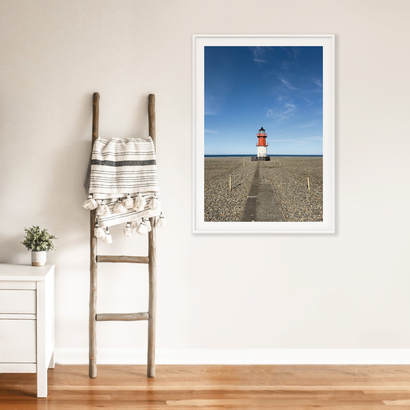 Vertical landscape photograph of a red and white striped lighthouse on tidal flats in a white frame with white mat, hung on a cream wall above a wooden ladder with rolled linens, styled with a small white side table displaying a potted plant on a warm wood floor.