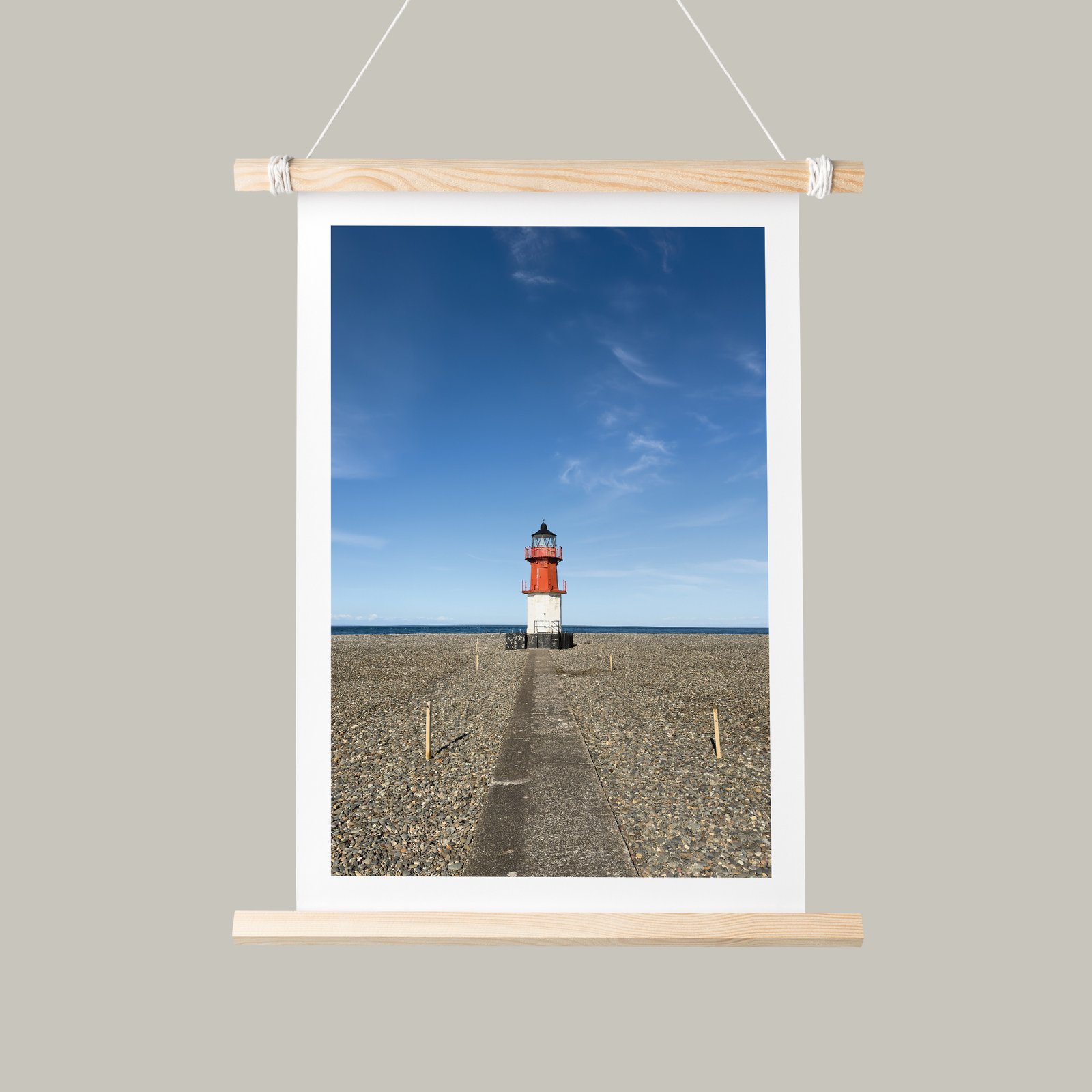 Vertical landscape photograph of a solitary lighthouse on mudflats beneath blue sky mounted as a hanging banner with white mat, natural wood dowel top and bottom frame with rope suspension, displayed against a soft sage-green background.