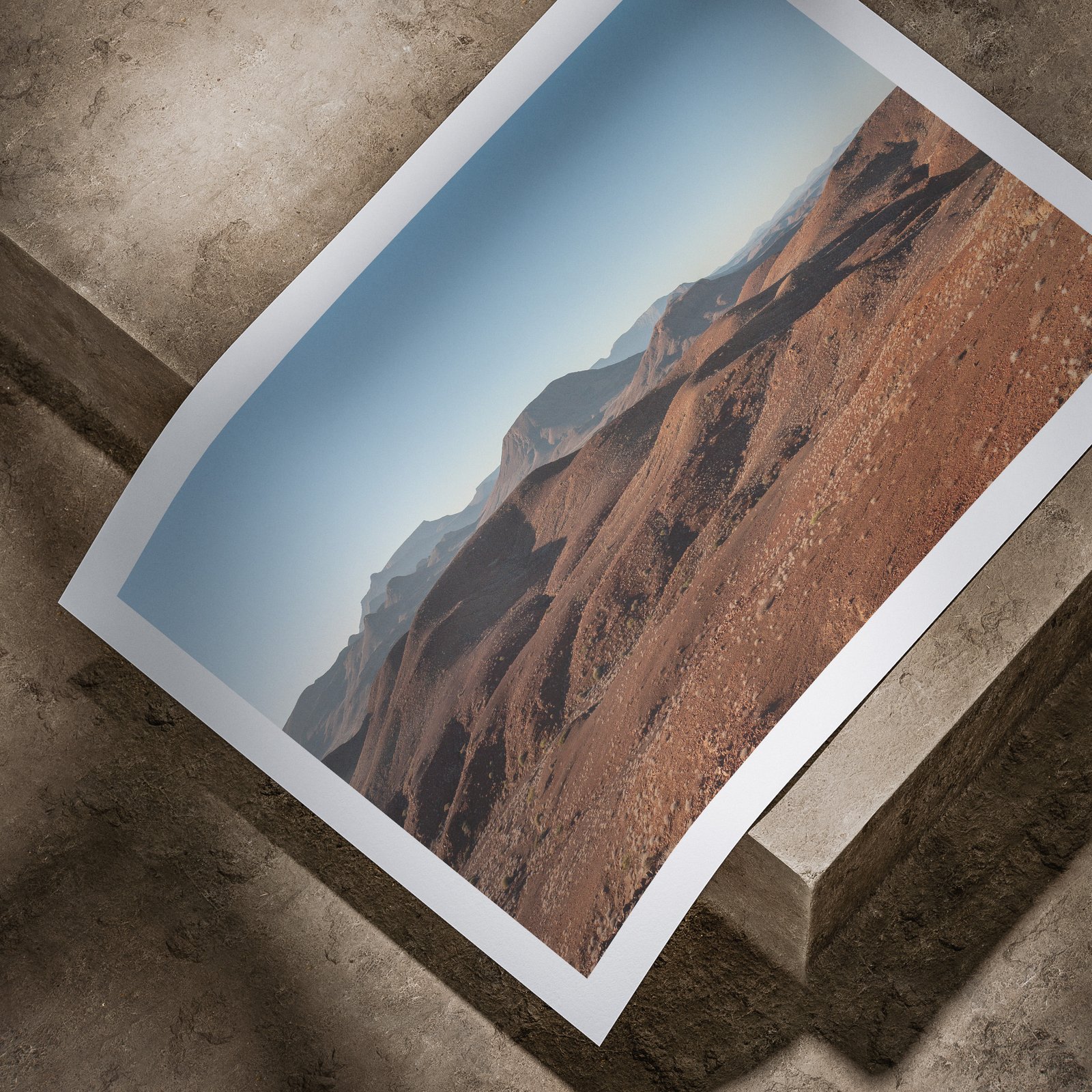 Horizontal landscape photograph of Namibian desert with volcanic peaks and layered terrain printed on white paper, laid flat on a concrete surface at an angled perspective.