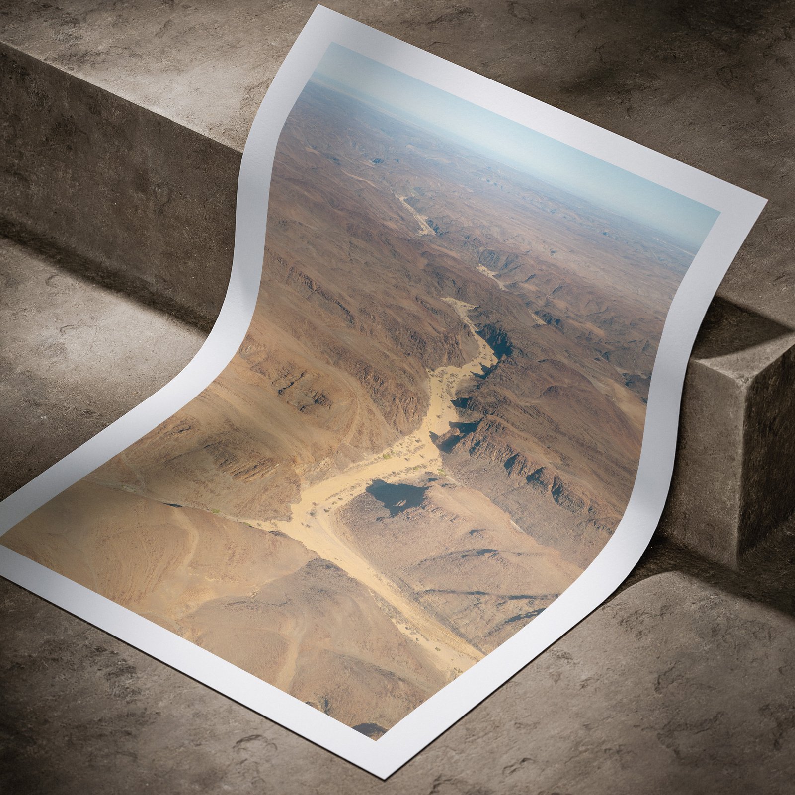Vertical aerial photograph of a meandering river through desert terrain printed on white paper, shown in a curved, rolled perspective on a concrete surface with dramatic lighting highlighting the print edges.