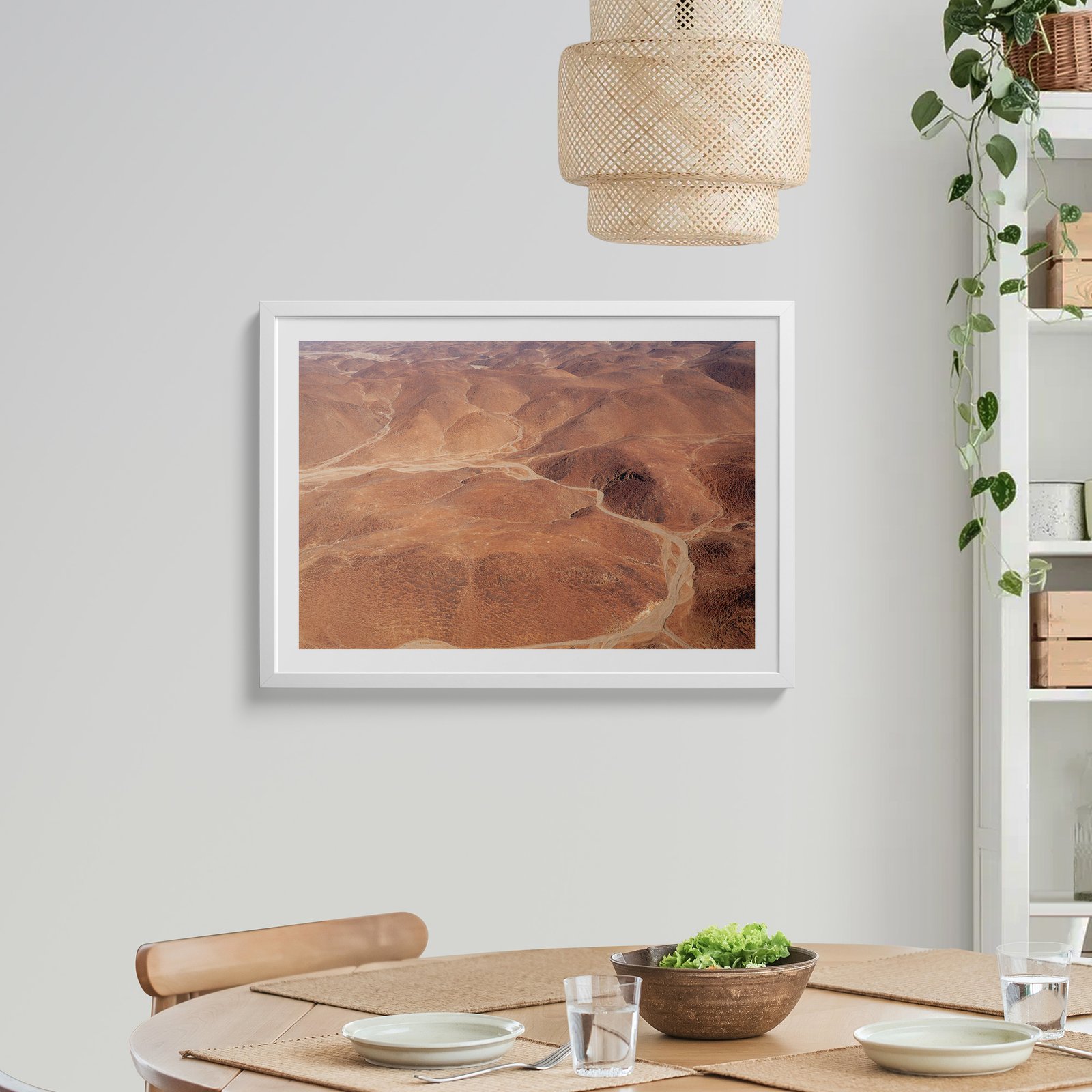 Horizontal aerial landscape photograph in a white frame with white mat, hung on a light wall above a wooden dining table, styled with a rattan pendant light above, trailing ivy plant to the side, and table settings below.