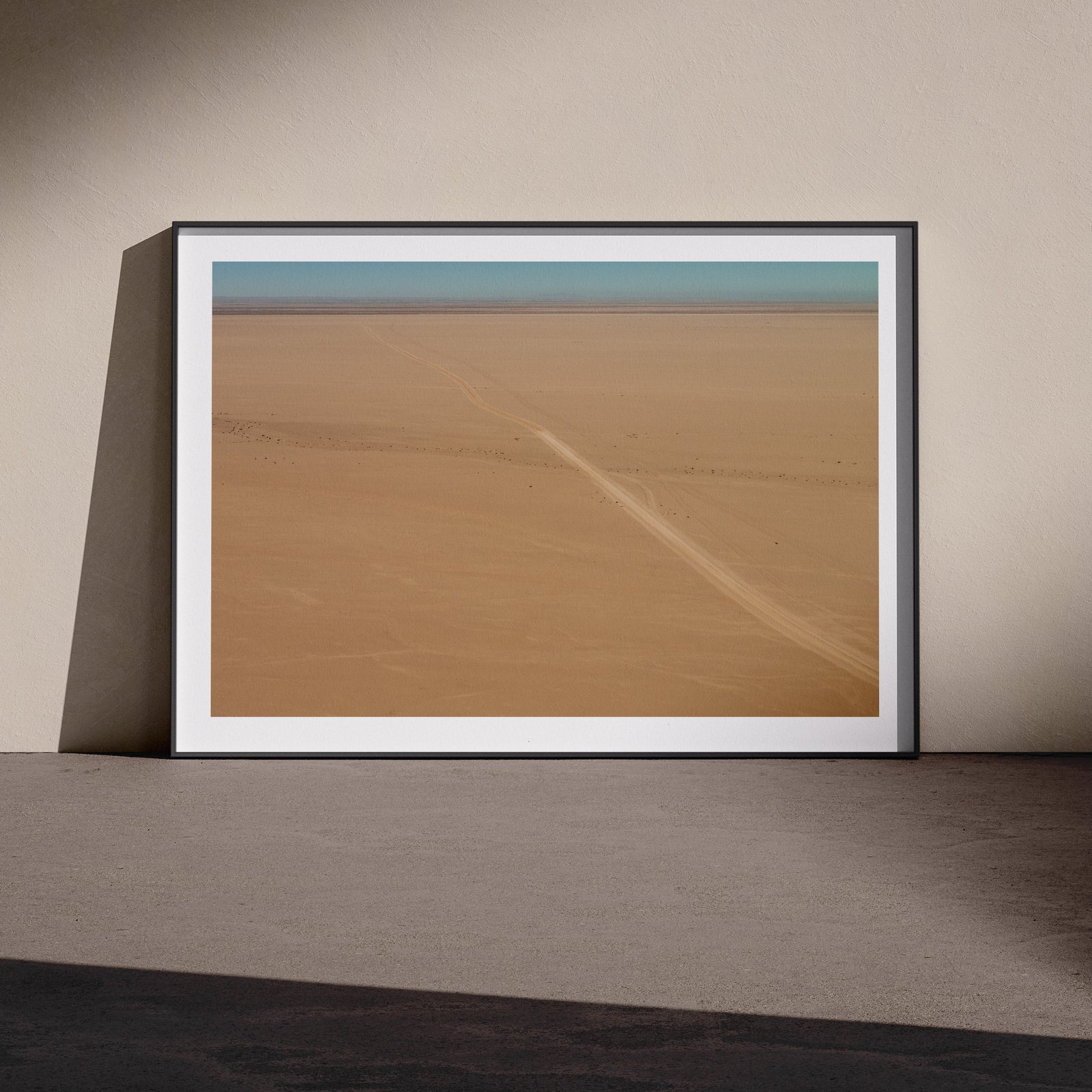 Horizontal landscape photograph with turquoise sky and tan desert sand in a white frame with subtle shadow mat, leaning against a pale wall in a gallery-style floor presentation, lit with soft studio lighting creating defined shadows.