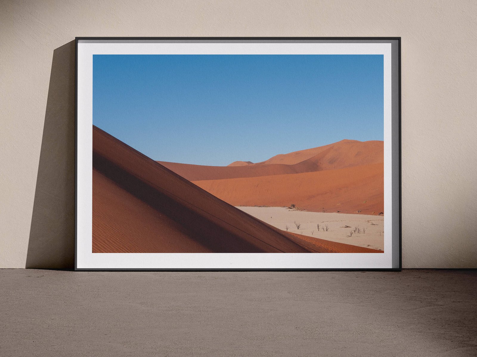 Horizontal landscape photograph of a Namibian sand dune with blue sky and warm sand tones in a white frame with dark shadow mat, leaning against a pale wall in a gallery-style floor presentation, lit with soft studio lighting creating defined shadows.