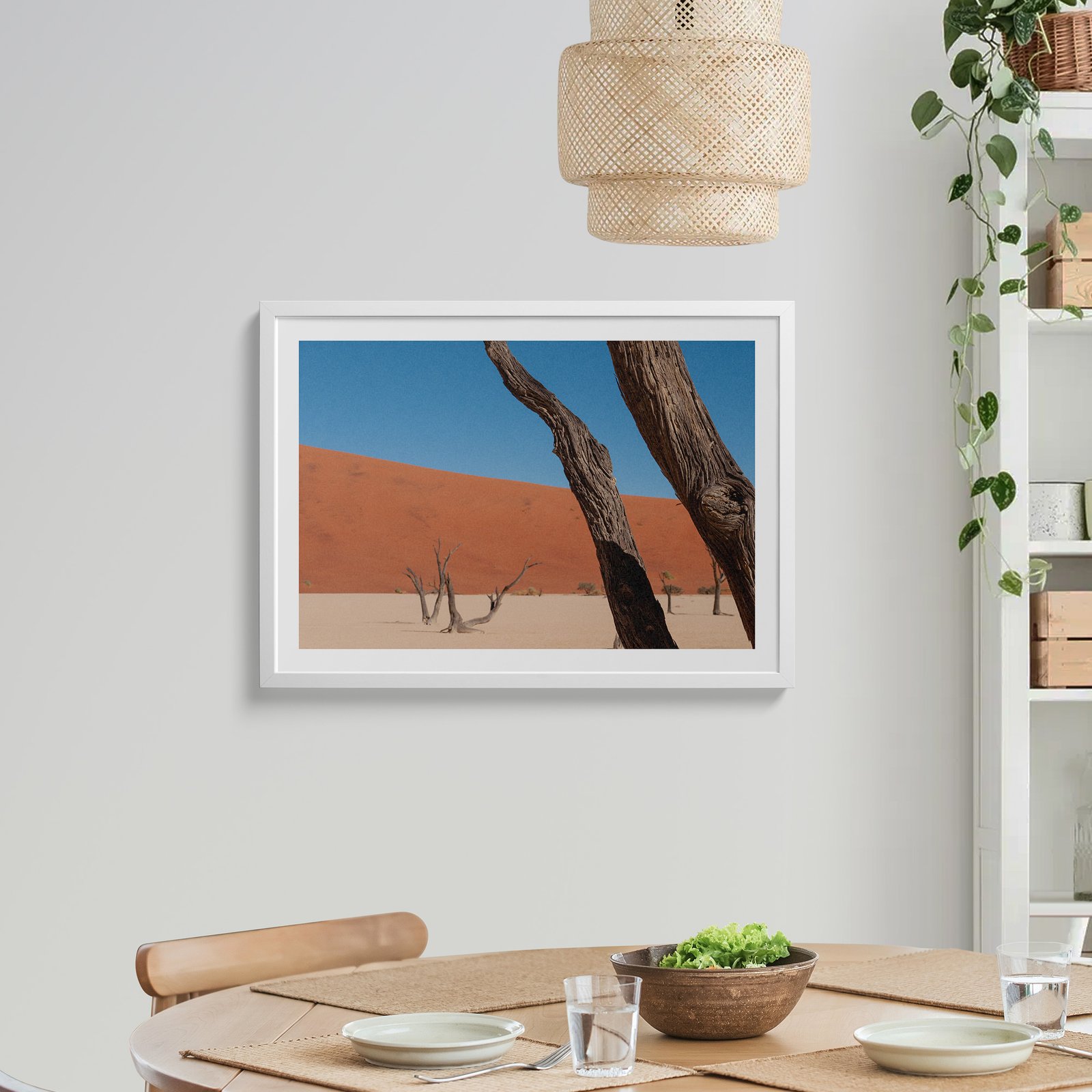Horizontal landscape photograph of Deadvlei dead trees silhouetted against orange dunes and clear sky in a white frame with white mat, hung on a light wall above a wooden dining table, styled with a rattan pendant light above, trailing ivy plant to the side, and place settings below.