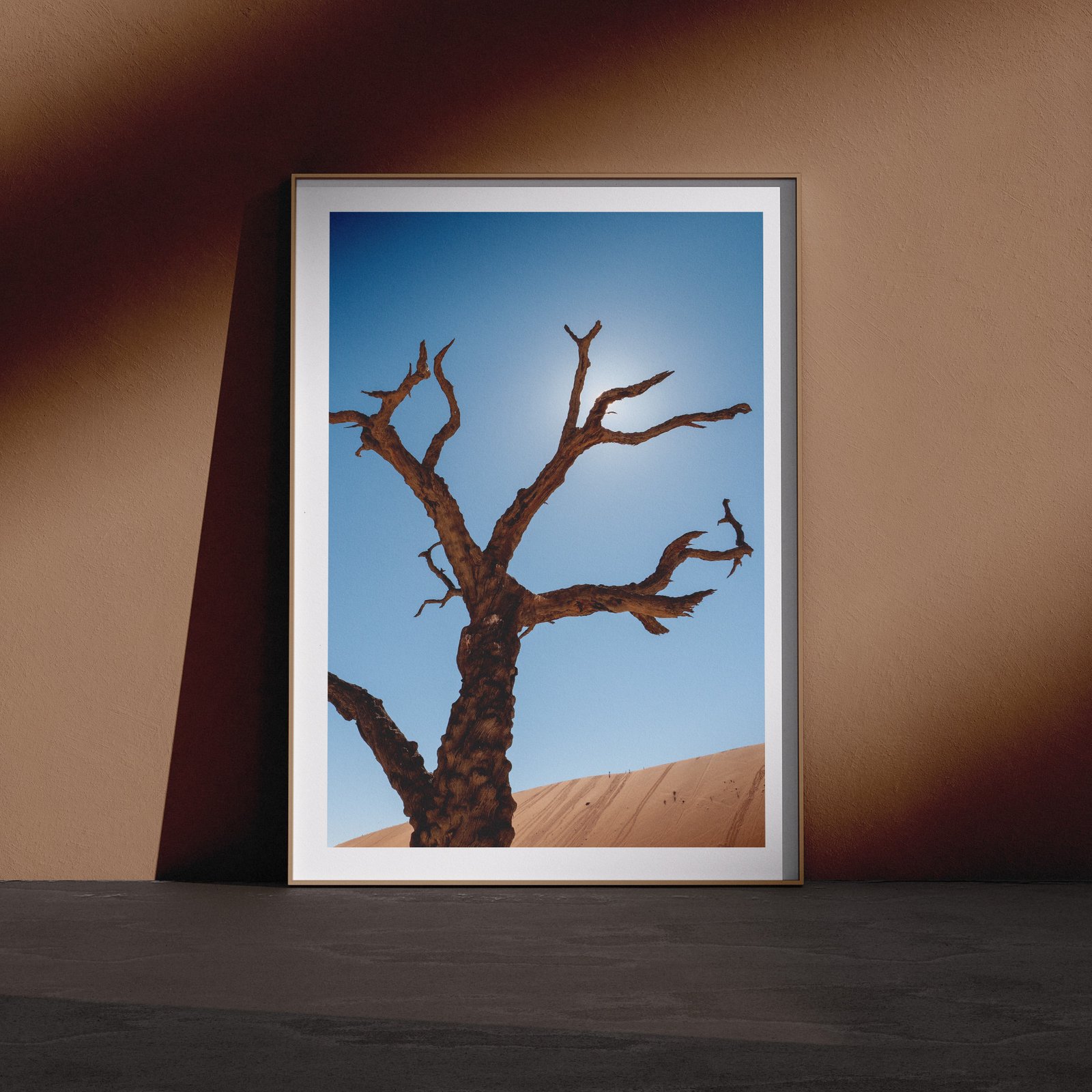 Vertical landscape photograph of a Deadvlei dead tree silhouetted against clear blue sky and sand dune in a white frame with white mat, leaning against a dark brown wall in a gallery-style floor presentation with dramatic side lighting.
