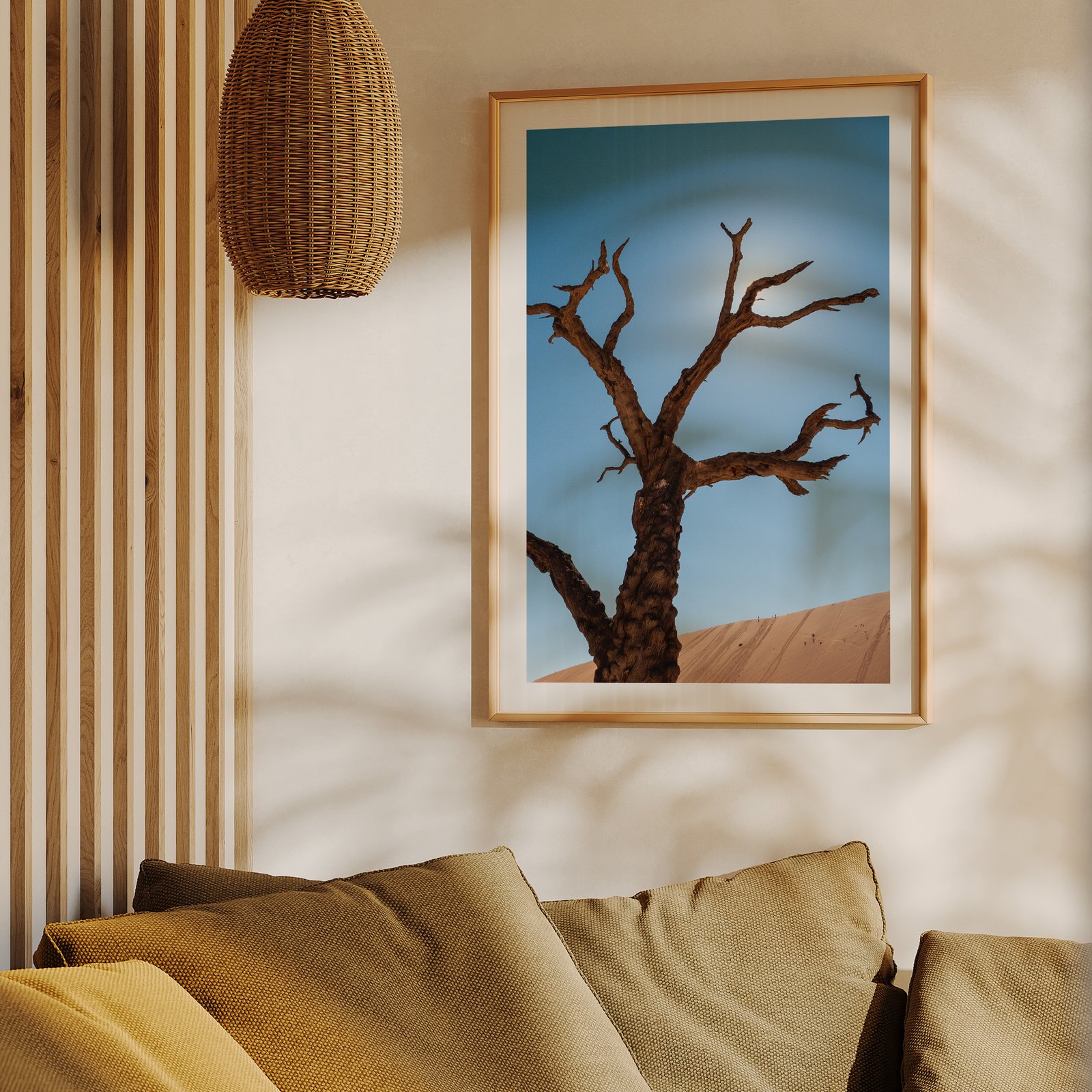 Vertical landscape photograph of a Deadvlei dead tree against blue sky and terracotta dune in a natural wood frame, hung on a cream wall above a mustard yellow upholstered sofa, styled with a woven rattan pendant light visible above.
