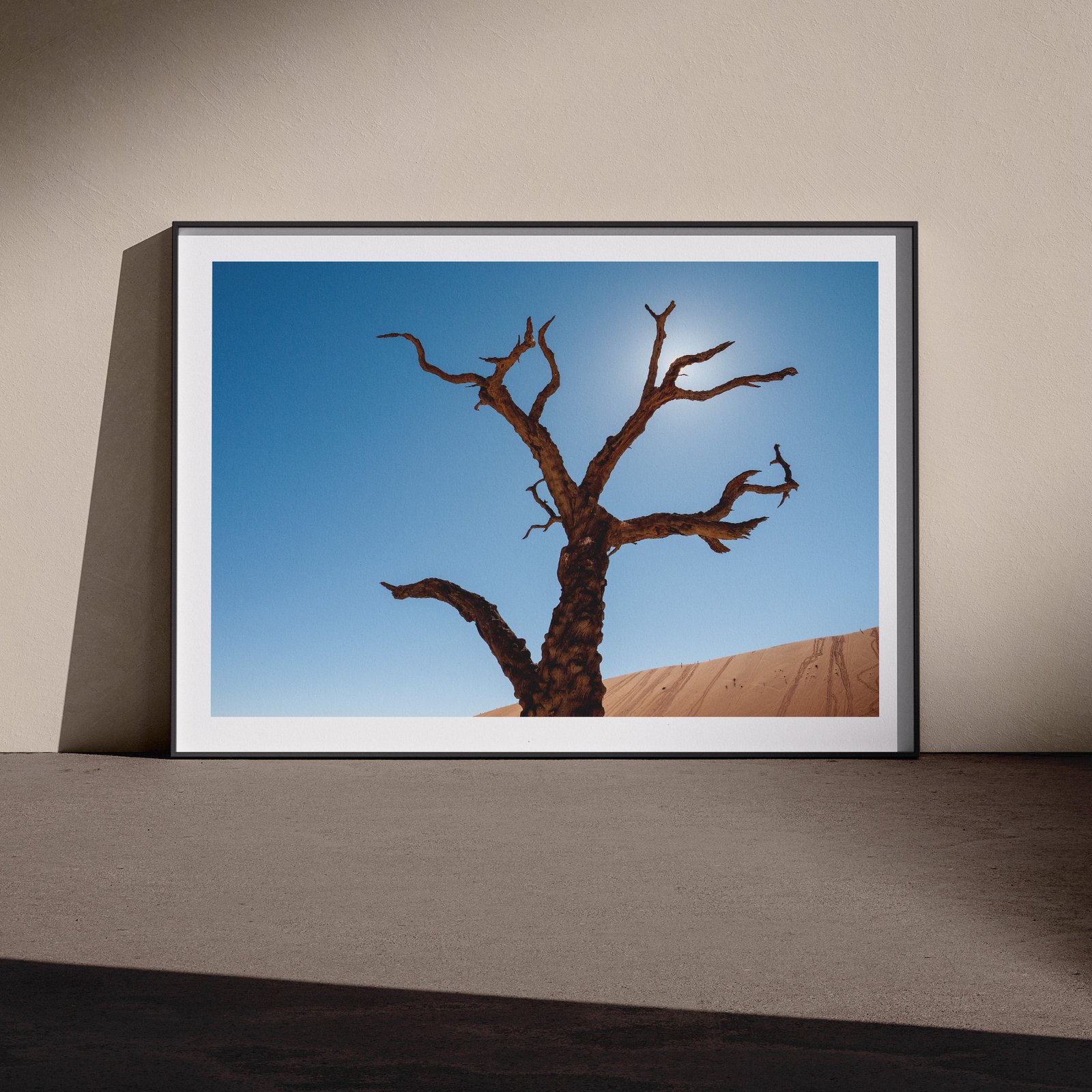 Vertical landscape photograph of a Deadvlei dead tree silhouetted against clear blue sky and terracotta dune in a white frame with dark shadow mat, leaning against a pale wall in a gallery-style floor presentation, lit with soft studio lighting.