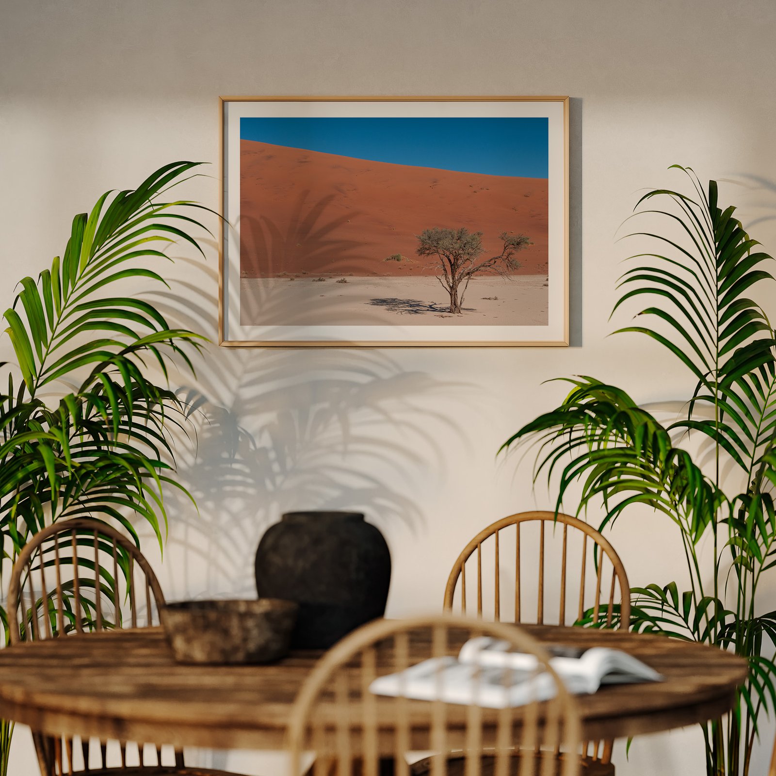 Horizontal landscape photograph of a solitary desert tree against blue sky and rust-colored dune in a natural wood frame with white mat, hung on a cream wall above a wooden dining table, styled with potted palm plants flanking the composition and dining chairs visible below.