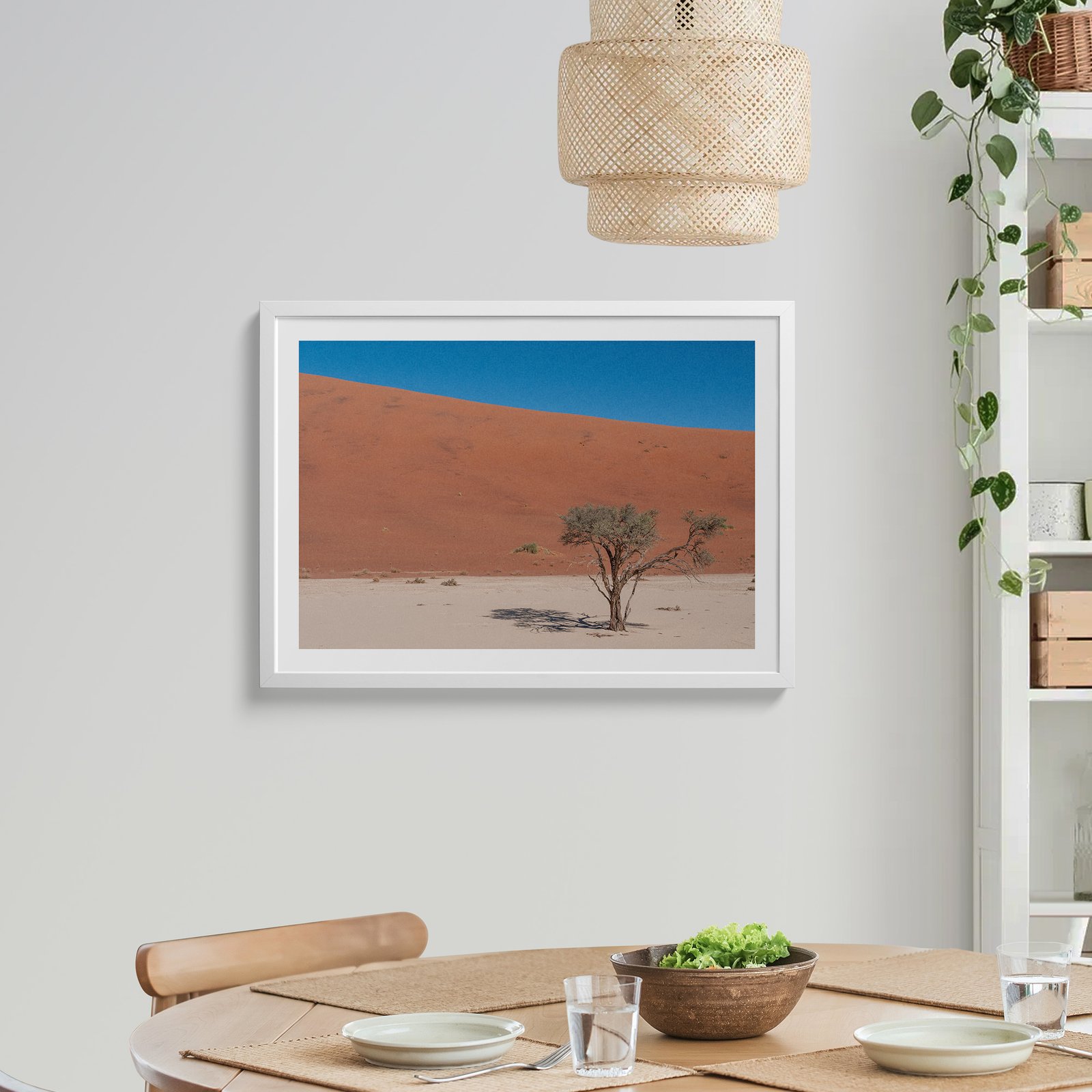 Horizontal landscape photograph of a solitary desert tree against terracotta dune and clear sky in a white frame with white mat, hung on a light wall above a wooden dining table, styled with a rattan pendant light above, trailing ivy plant to the side, and place settings below.