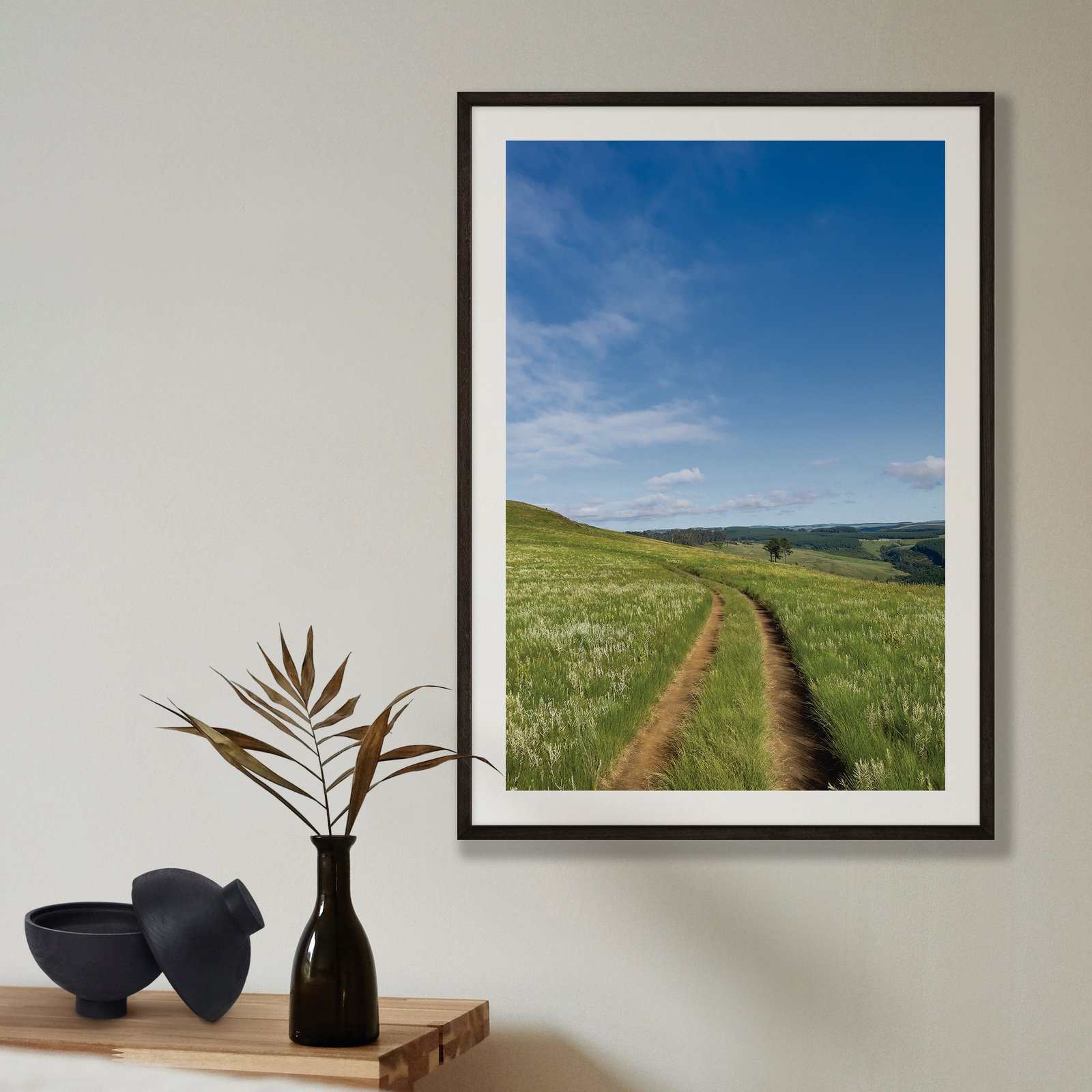 Vertical landscape photograph of a worn track winding through lush green fields beneath a clear blue sky, displayed in a black frame with white mat on a pale beige wall, styled with a minimalist shelf arrangement below featuring a black vase with dried grasses and black ceramic bowl.