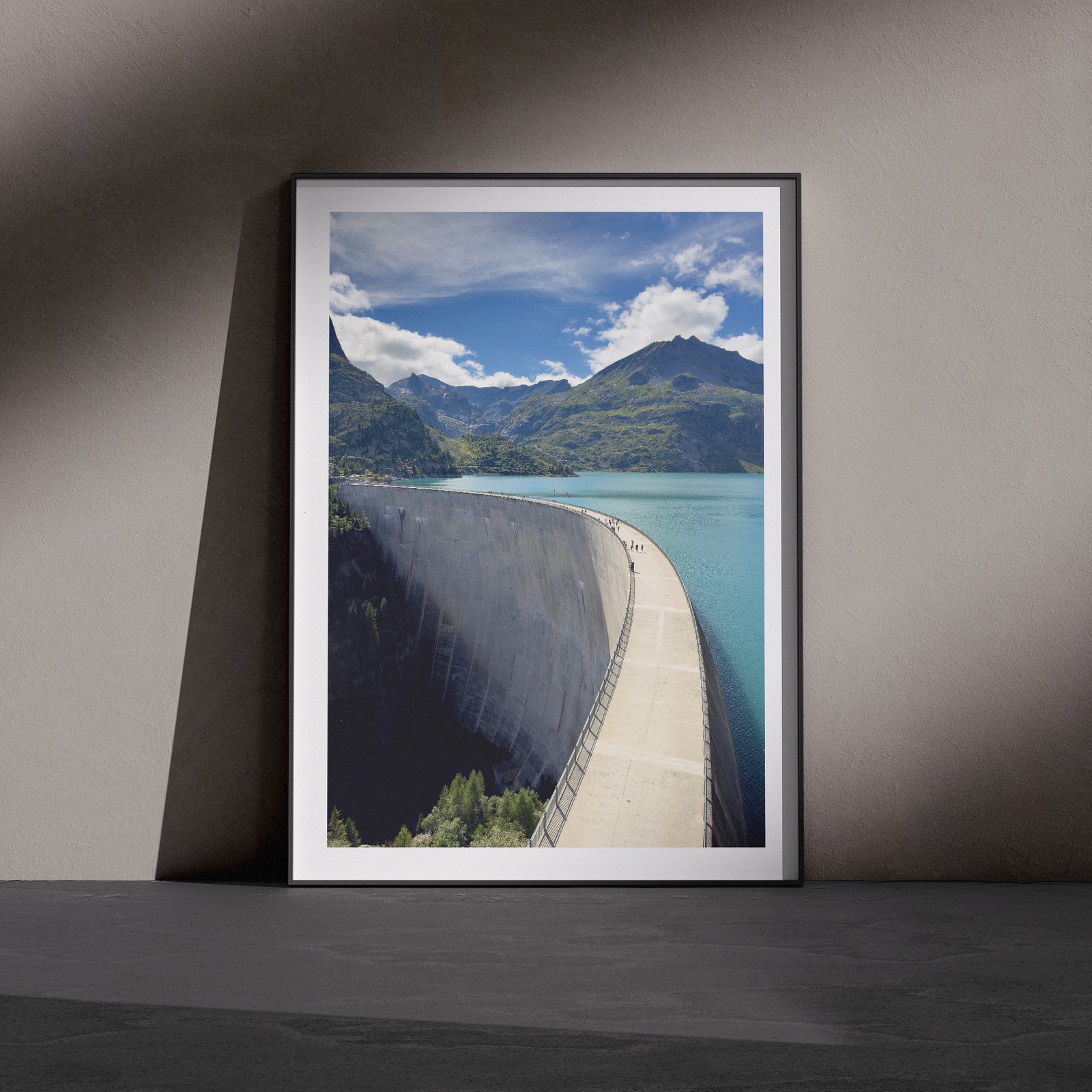 Vertical landscape photograph of a dam structure with turquoise water, mountain peaks, and clear blue sky in a white frame with white mat, leaning against a dark charcoal wall in a gallery-style floor presentation with dramatic side lighting creating defined shadows.