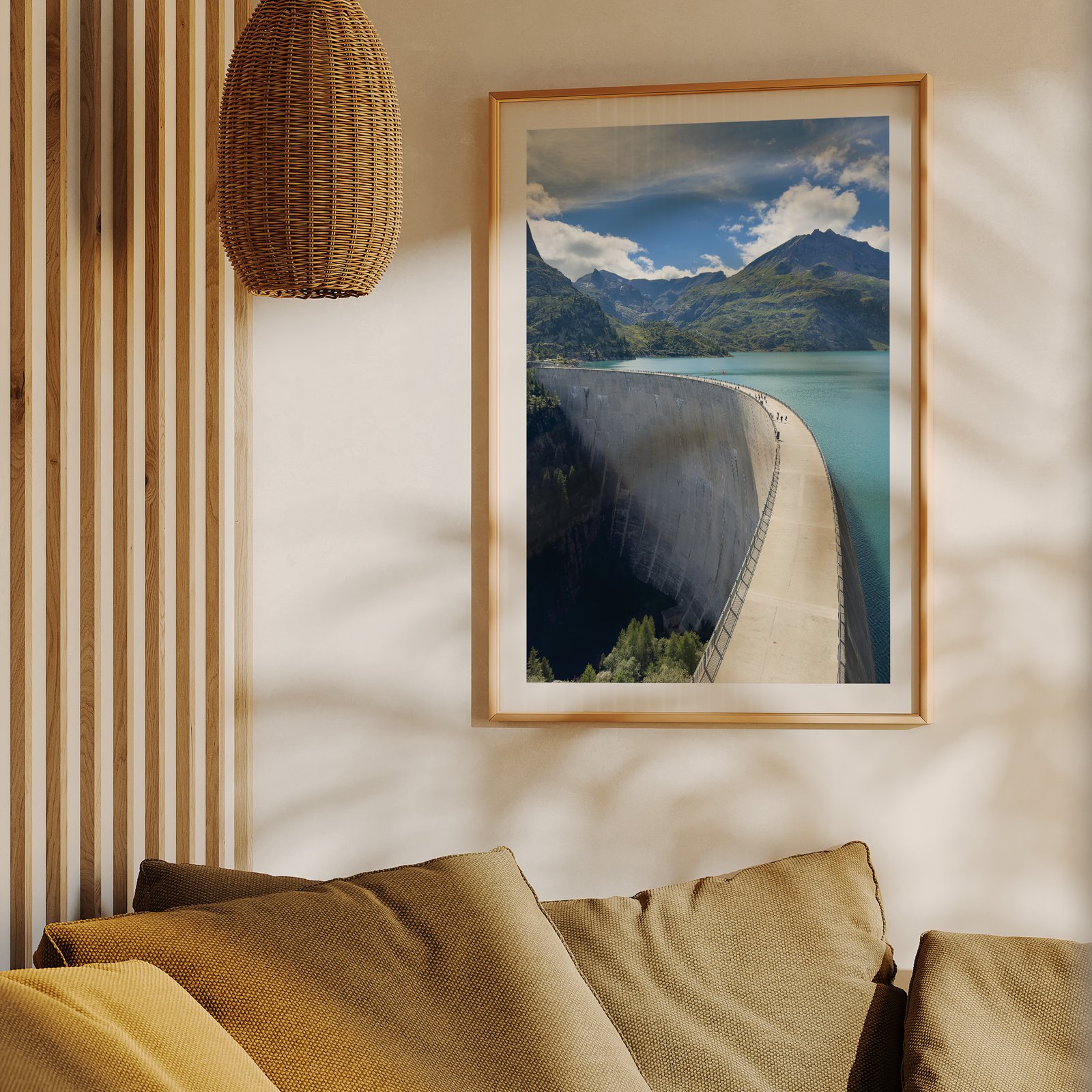Vertical landscape photograph of a concrete dam with turquoise alpine lake, forested shoreline, and snow-capped mountains under blue sky, displayed in a natural wood frame hung on a cream wall above a mustard-gold upholstered sofa, styled with a woven rattan pendant light above and wooden slat wall paneling visible to the side.