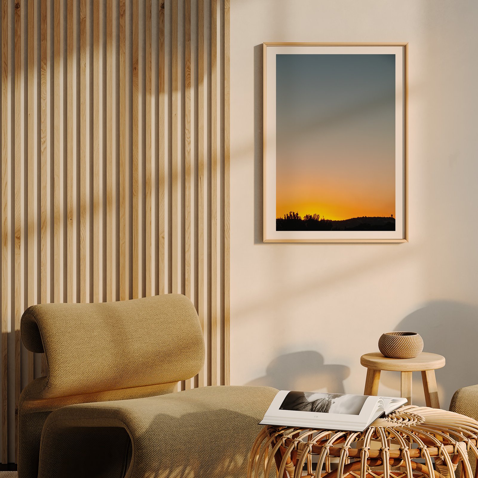 Vertical landscape photograph of a gradient sunset sky and silhouetted landscape in a natural wood frame, hung on a cream wall with wooden slat paneling visible to the side, above a golden-yellow upholstered chair with a woven wooden side table and magazines displayed below.