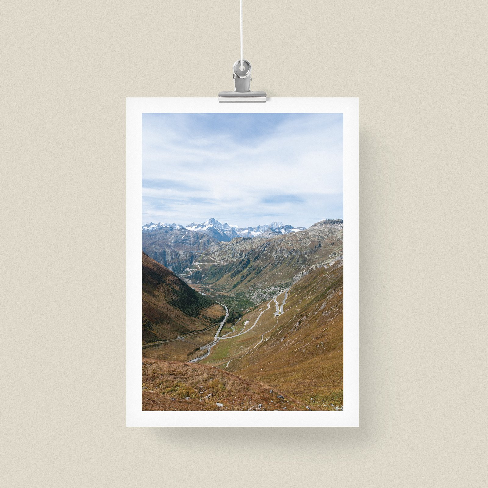 Vertical landscape photograph of a mountain valley with distant snow peaks and brown moorland in a white frame with white mat, suspended from ceiling with a metal clip-hanging mechanism against a pale cream background.
