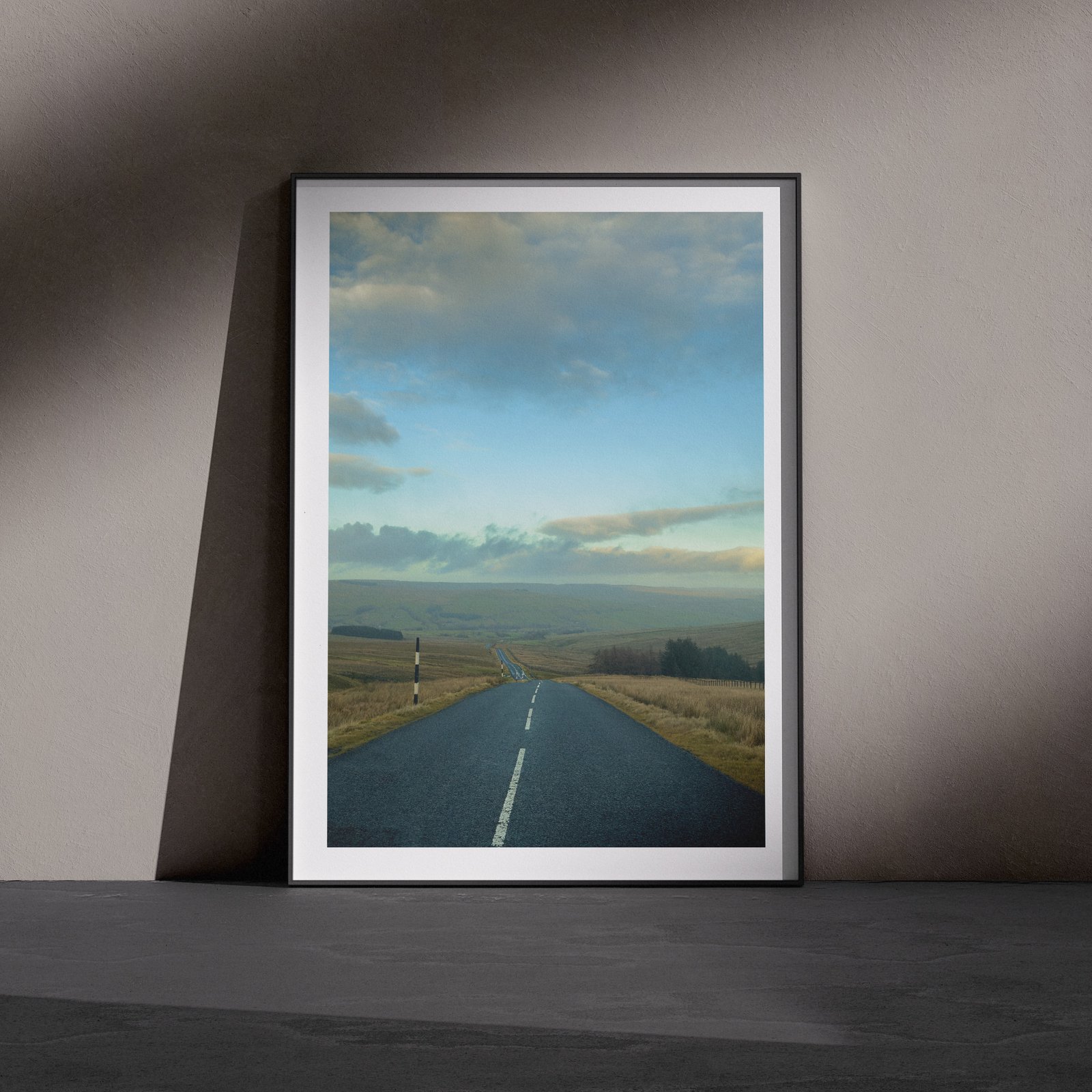 Vertical landscape photograph of a straight asphalt road stretching through moorland beneath a cloudy blue sky, displayed in a white frame with white mat, leaning against a dark charcoal wall in a gallery-style floor presentation with dramatic side lighting.