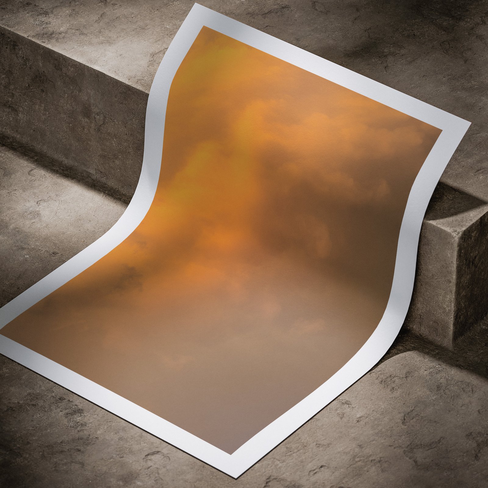 Vertical abstract photograph with golden orange and brown gradient tones printed on white paper, displayed in a curved, rolled perspective on a concrete surface with dramatic angled lighting highlighting the print edges and subtle color depth.