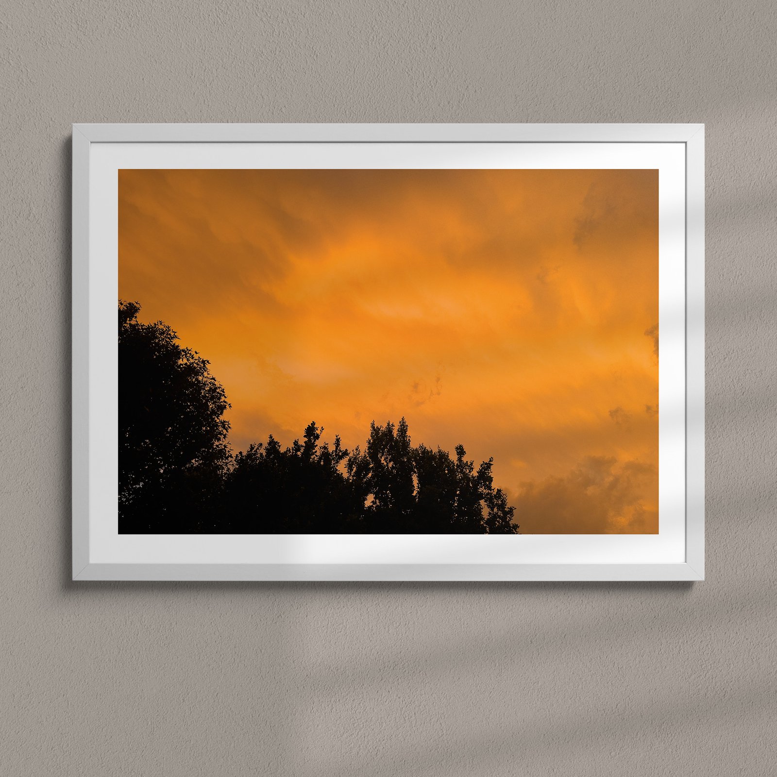 Horizontal landscape photograph of a warm golden-orange sunset with dark silhouetted forest below in a white frame with white mat, hung on a soft gray wall, lit with soft studio lighting creating subtle depth and shadows.