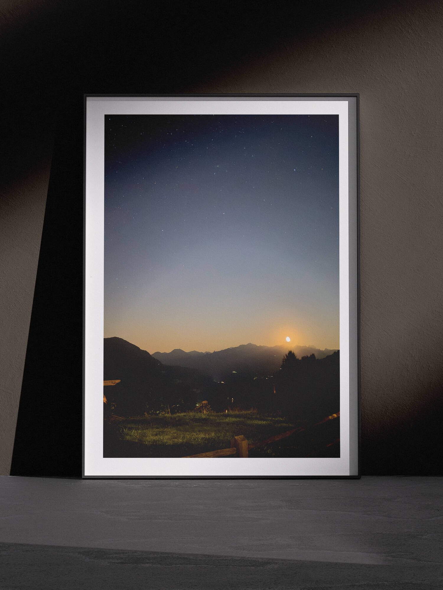 Horizontal landscape photograph of a sunrise or sunset over a distant mountain valley with forested slopes in a white frame, leaning against a dark charcoal wall in a gallery-style floor presentation with dramatic side lighting.