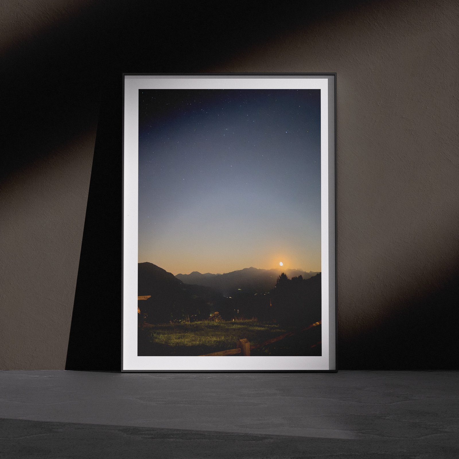 Horizontal landscape photograph of a sunrise or sunset over a distant mountain valley with forested slopes in a white frame, leaning against a dark charcoal wall in a gallery-style floor presentation with dramatic side lighting.