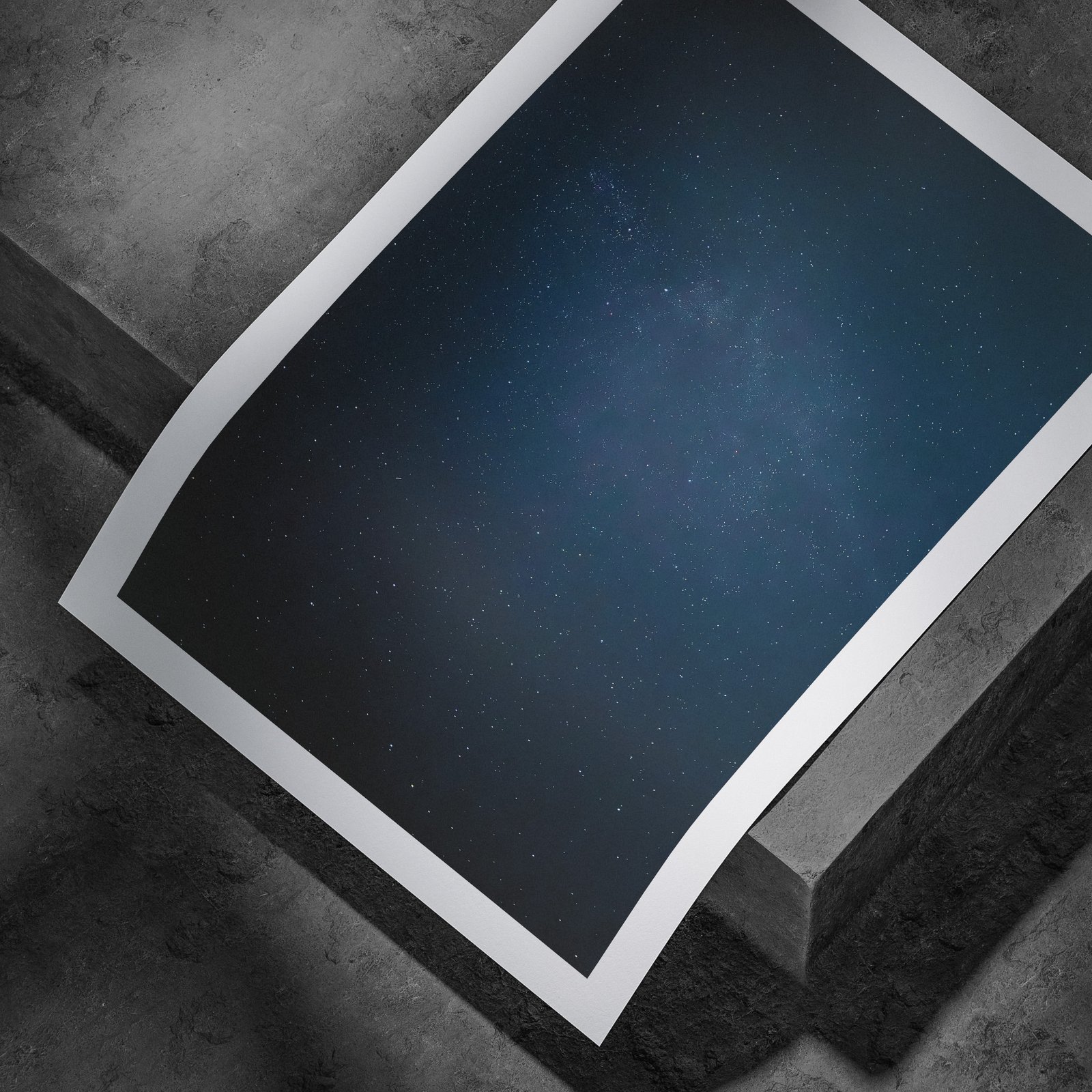 Horizontal starfield photograph with deep blue night sky and visible stars printed on white paper, displayed in a curved, rolled perspective on a concrete surface with dramatic angled lighting highlighting the print edges.