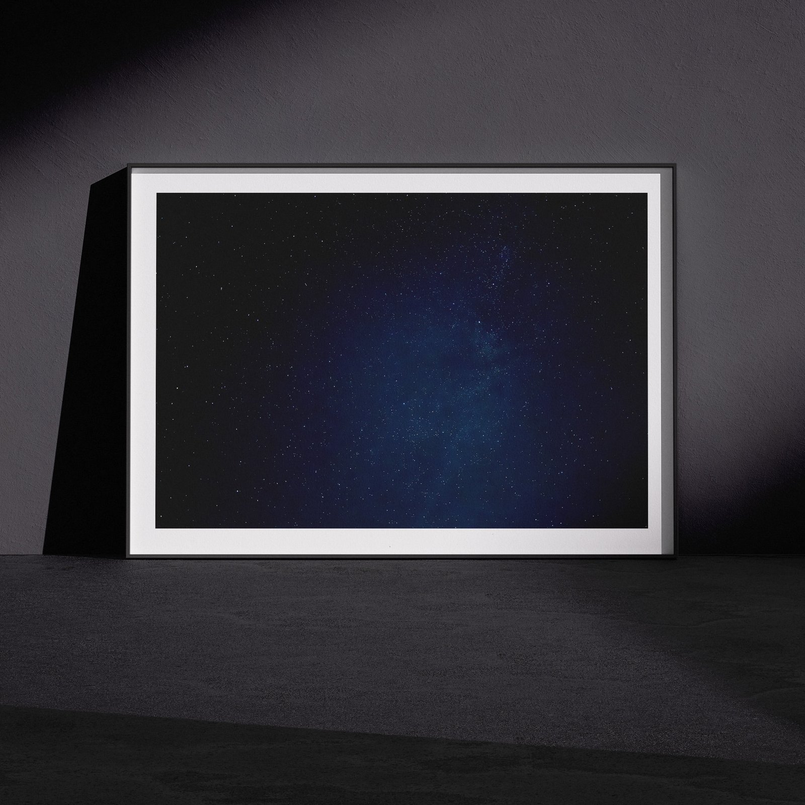 Horizontal starfield photograph with deep blue night sky in a white frame, leaning against a dark charcoal wall in a gallery-style floor presentation with dramatic side lighting creating subtle shadows.