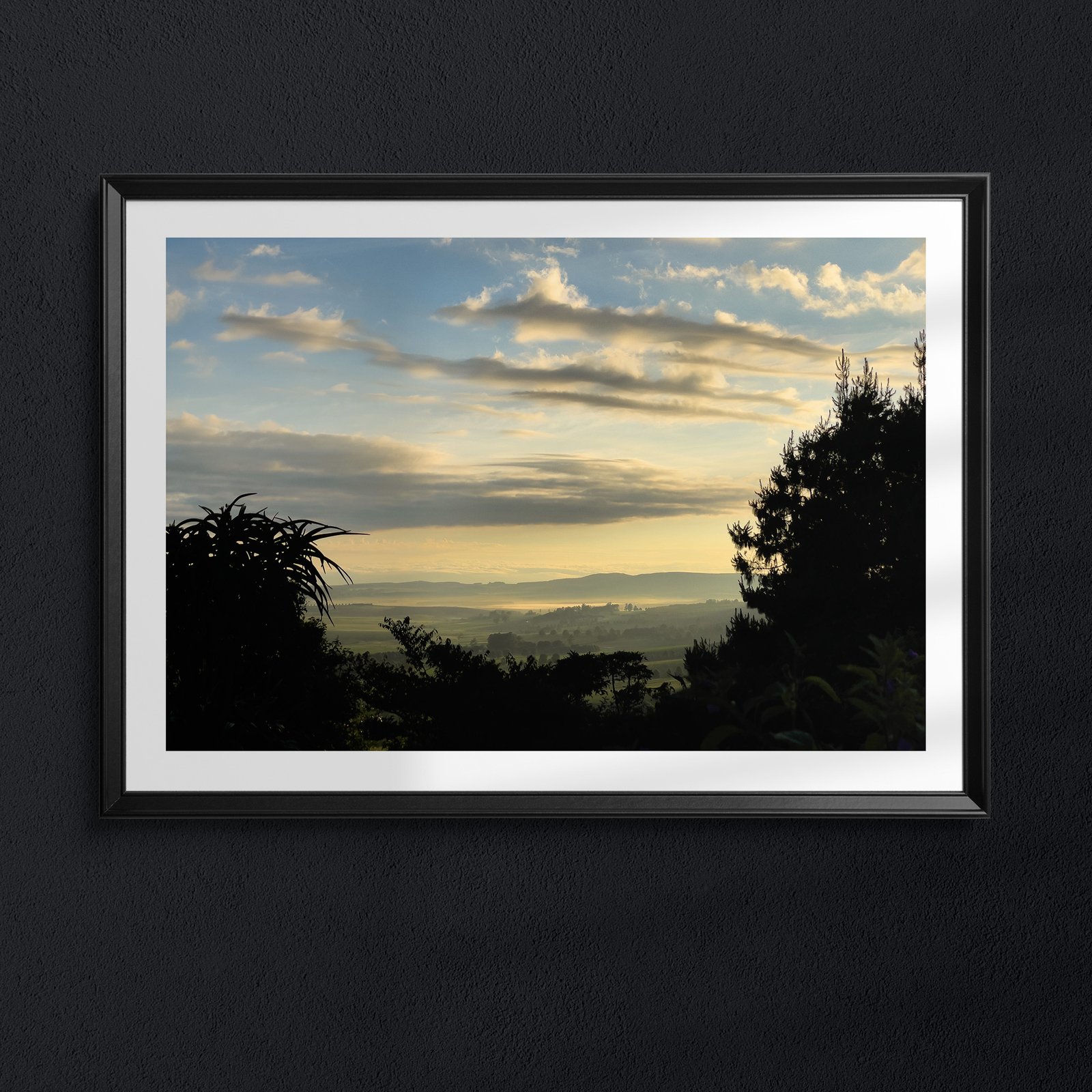 Horizontal landscape photograph of a golden-hour sunset with warm cream and blue tones, silhouetted tropical vegetation and palm fronds in the foreground, displayed in a white frame with black outer frame on a dark charcoal wall, creating dramatic high-contrast presentation.