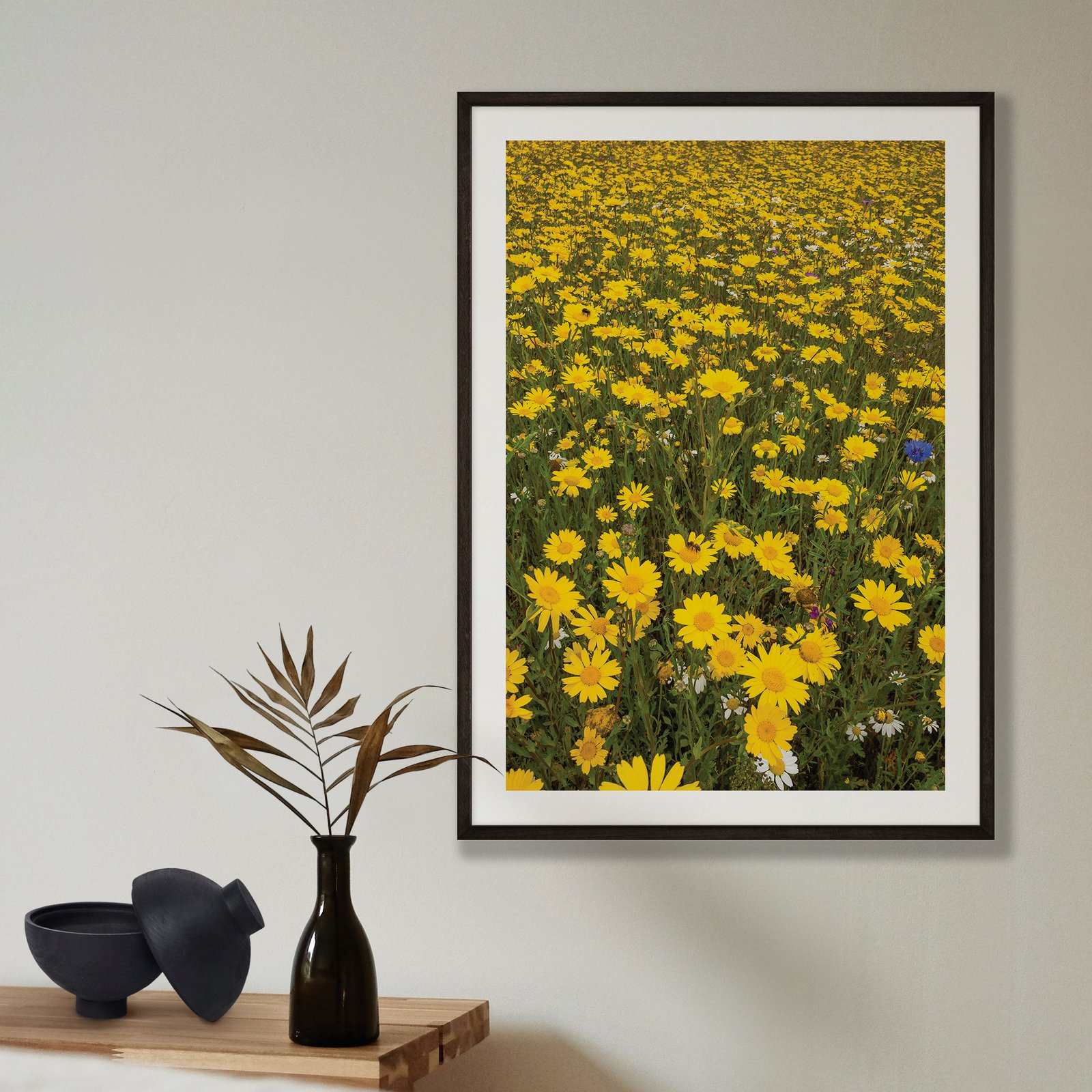 Vertical landscape photograph of a dense field of vibrant yellow daisies or wildflowers with green foliage, displayed in a black frame with white mat on a pale beige wall, styled with a minimalist shelf arrangement below featuring a black vase with dried grasses and black ceramic bowl.